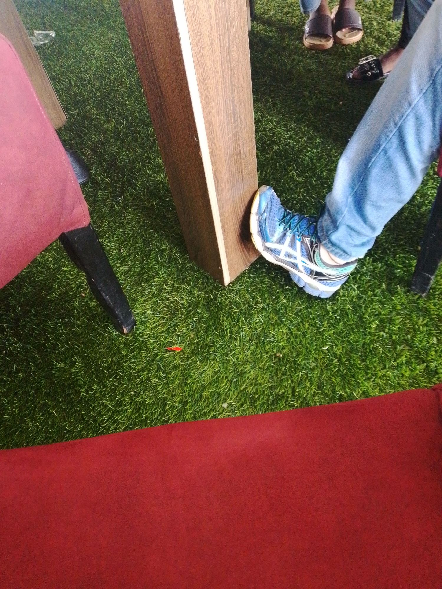 The image captures a close-up view of a person's legs and feet on artificial grass, near a wooden table leg and a red chair. The person is wearing blue jeans and blue and white athletic shoes. In the background, two other individuals are partially visible, one wearing dark sandals and the other wearing black sandals. The setting appears to be an outdoor or indoor area with a faux grass flooring, possibly a cafe or recreational space. There is no information about the location being in Barikin Jirga Summa, Nigeria, within the provided image.