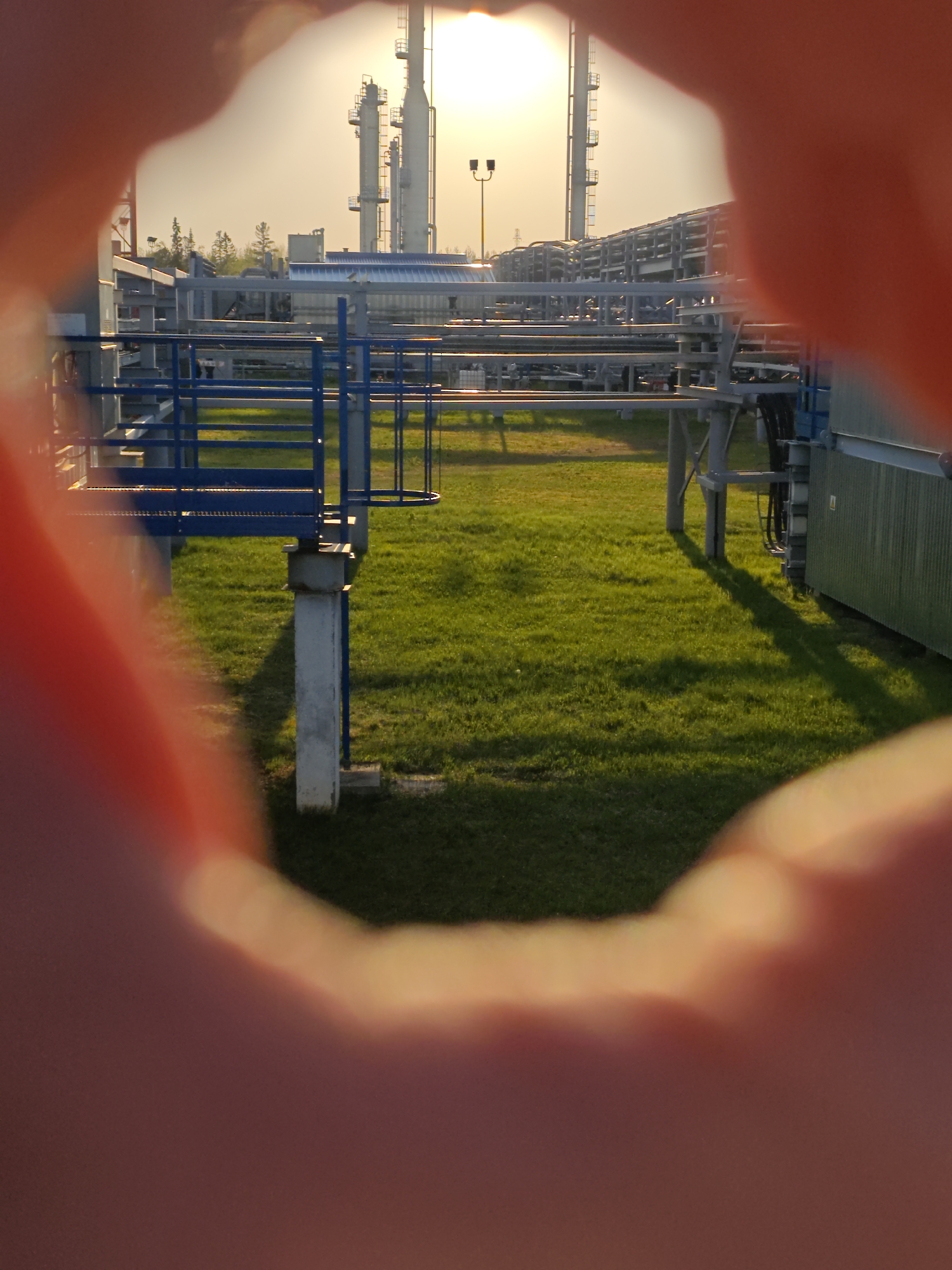 The image depicts an industrial landscape in what appears to be Unknown, Russia, framed by a blurred foreground suggesting it is being viewed through a hand-made "scope". The mid-ground shows a patch of vibrant green grass, partially shadowed, upon which stand sections of metal pipes and infrastructure, painted primarily in silver and blue. These structures include walkways with blue railings and supports of various sizes. In the background, refinery towers rise against a hazy, sunlit sky. A street light pole is also visible. The lighting suggests either sunrise or sunset.