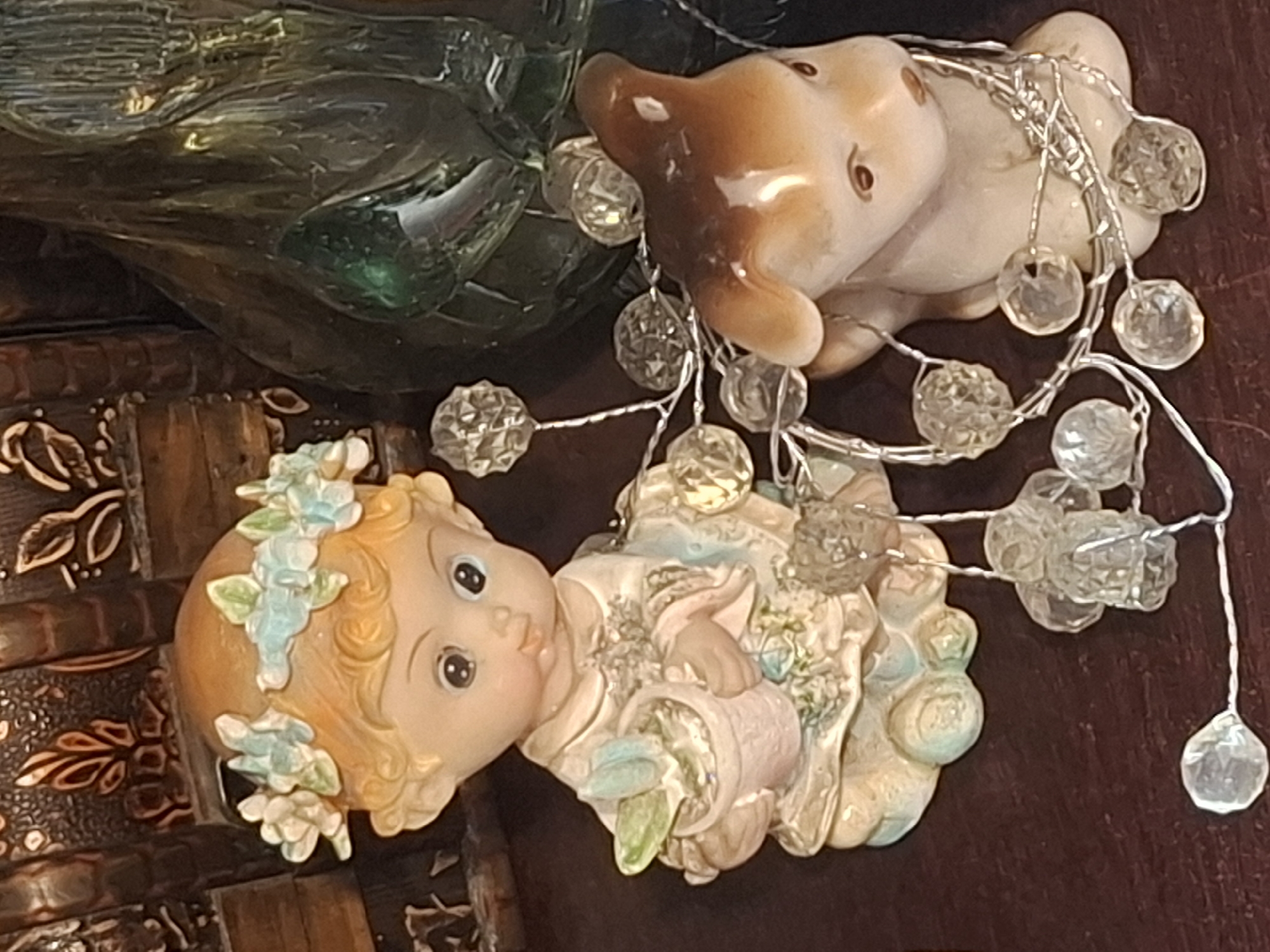 This is a close-up, eye-level, indoor photograph of two small decorative figurines on a dark brown surface. On the left is a figurine of a child with blonde hair, wearing a floral crown and a light-colored, patterned garment. The child holds a small pink pot with a green plant. The child's expression is wide-eyed and slightly surprised. To the right of the child, and slightly behind it, is a figurine of a brown and white rabbit. Both figurines are partially wrapped in a strand of small, clear, faceted decorative beads on a thin silver wire, which resembles a string of fairy lights. In the background, to the left, is a dark, ornate, possibly wooden object with raised decorative elements. To the right of the child figurine and behind the rabbit, there is a dark, translucent, green object that appears to be glass. The lighting is warm and appears to be artificial, casting soft shadows. There is no discernible connection to the city of Almaty, Kazakhstan.