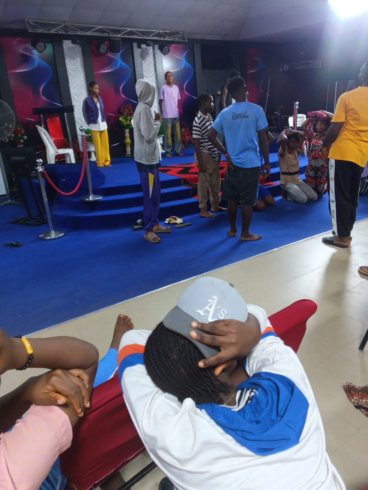 The image depicts an indoor gathering, possibly a church service or event, set in Nkpor, Nigeria. The scene is dominated by a raised platform with a blue carpeted stage, adorned with patterned backdrops featuring flowing blue and pink designs. Several individuals are present on the platform and in front of it.
On the platform, from left to right, a woman in a purple jacket and yellow pants stands facing the audience. Next to her, a person in a grey hoodie and purple pants is seen from the back. Further to the right, a young man in a pink t-shirt and patterned pants stands. A blue banner that reads "EZINNE" is visible on the t-shirt of a young man in the foreground. Another young man in a blue t-shirt and dark shorts is also visible. In the background, a woman in a colorful patterned dress is kneeling, with another person beside her. A person in a yellow t-shirt stands on the far right.
In the foreground, a person is seated, resting their head on their hands, wearing a white hoodie with blue accents and a grey baseball cap. Their legs are crossed, and they are wearing blue and orange striped pants. Another person's arm, wearing a pink sleeve and a yellow bracelet, is visible to the left. The floor in the foreground is light-colored, transitioning to the blue carpeted area.
Various objects are present, including white chairs on the platform, red velvet ropes with silver stanchions for crowd control, and decorative flowers in vases. Stage lighting fixtures are mounted above. The overall impression is of a communal gathering with a focus on the events taking place on the elevated platform.
