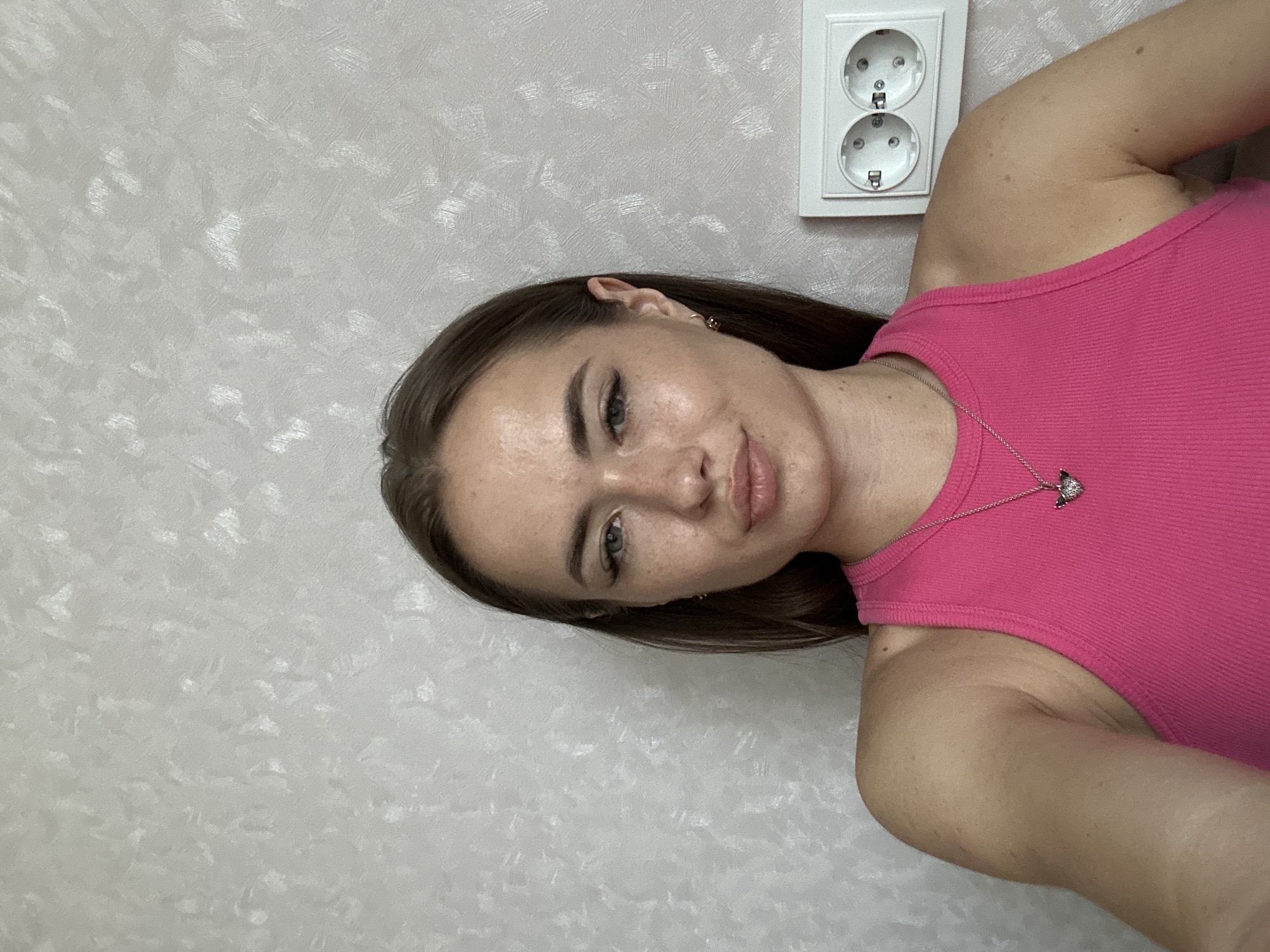 This media file is a portrait of a young woman, taken from a slightly high angle, indoors. The woman is facing the camera, with her head tilted to her right and her body turned to her left. She has long, dark brown hair and fair skin with freckles. She is wearing a bright pink, ribbed tank top and a delicate silver necklace with a small, heart-shaped pendant. Her expression is relaxed and natural, with a slight smile.
The background is a textured, light-colored wall, which appears to be wallpapered. To the right of the woman's head, mounted on the wall, is a white electrical outlet with two circular sockets.
The lighting appears to be soft and even, suggesting an indoor setting with artificial light or diffused natural light. There is no visible text in the image. The overall impression is that of a casual, personal photograph.
