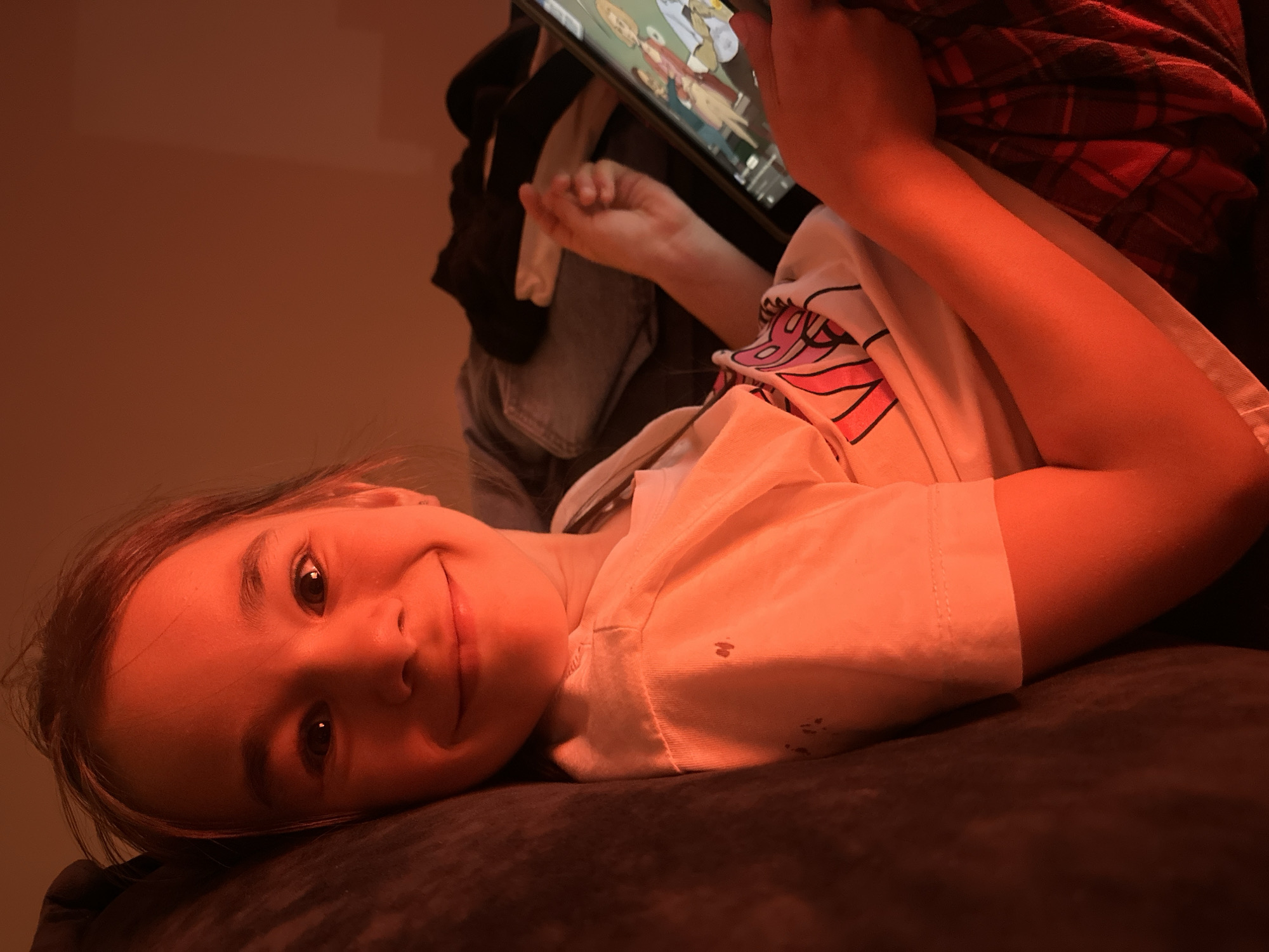 A child lies on a dark, plush surface, possibly a couch or bed. They are holding a tablet displaying cartoon figures. The child is smiling at the viewer. They are wearing a light-colored shirt with a pink and black design. There is clothing piled in the background, including a denim jacket, a black garment, and a red plaid flannel shirt. The lighting is warm, possibly from a lamp. The location of Kharnavy, Russia is not discernible in this image.