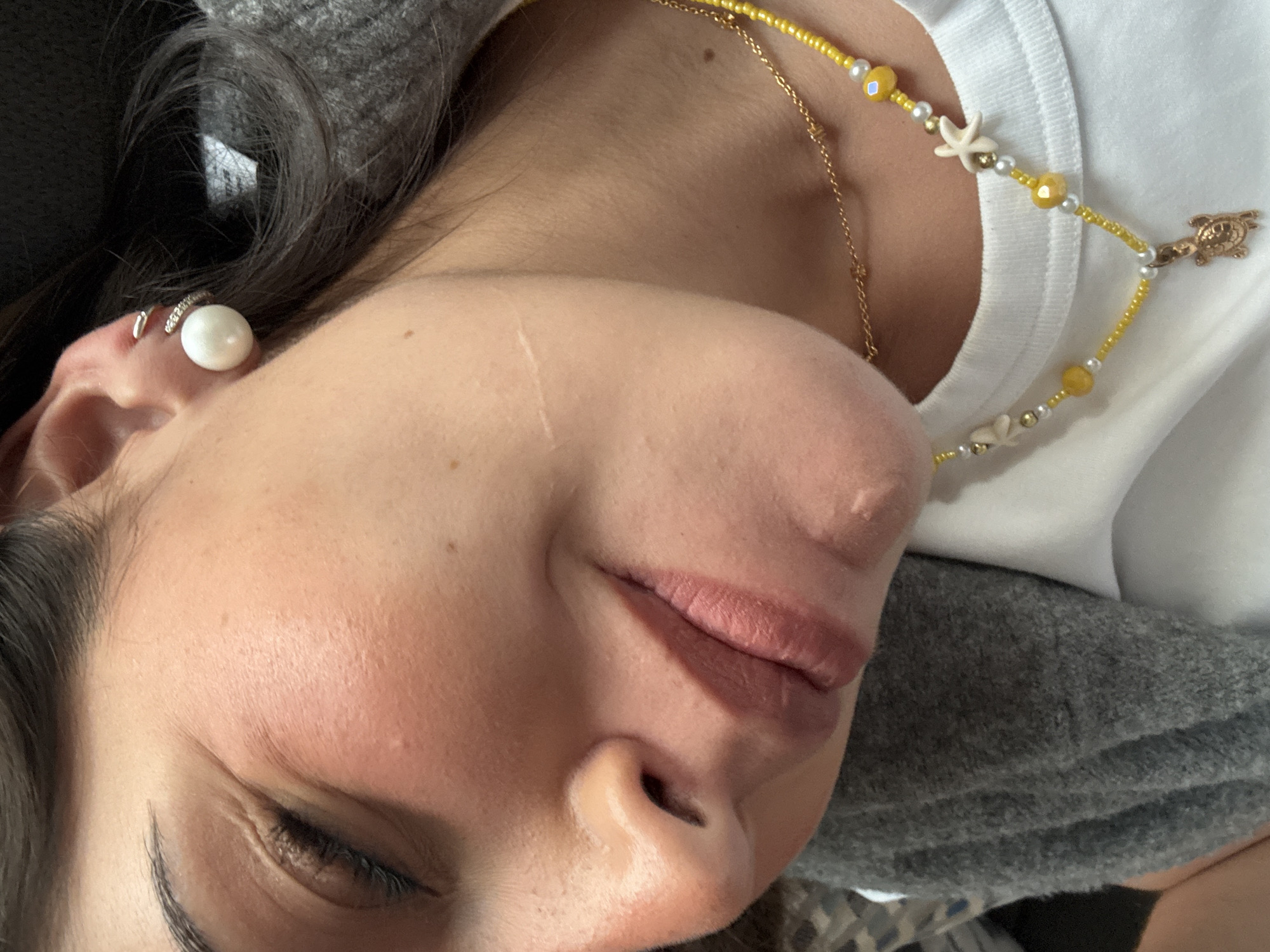 A close-up, angled head-and-shoulders shot of a person with light skin, closed eyes, and dark hair. The person is wearing a white t-shirt and a gray sweater. The person has a pearl earring and a beaded necklace with yellow beads, white starfish, and a turtle charm. A thin gold chain necklace is also visible. The location is not discernible from the image, and therefore information about San Bernardino Tlaxcalancingo, Mexico cannot be provided.
