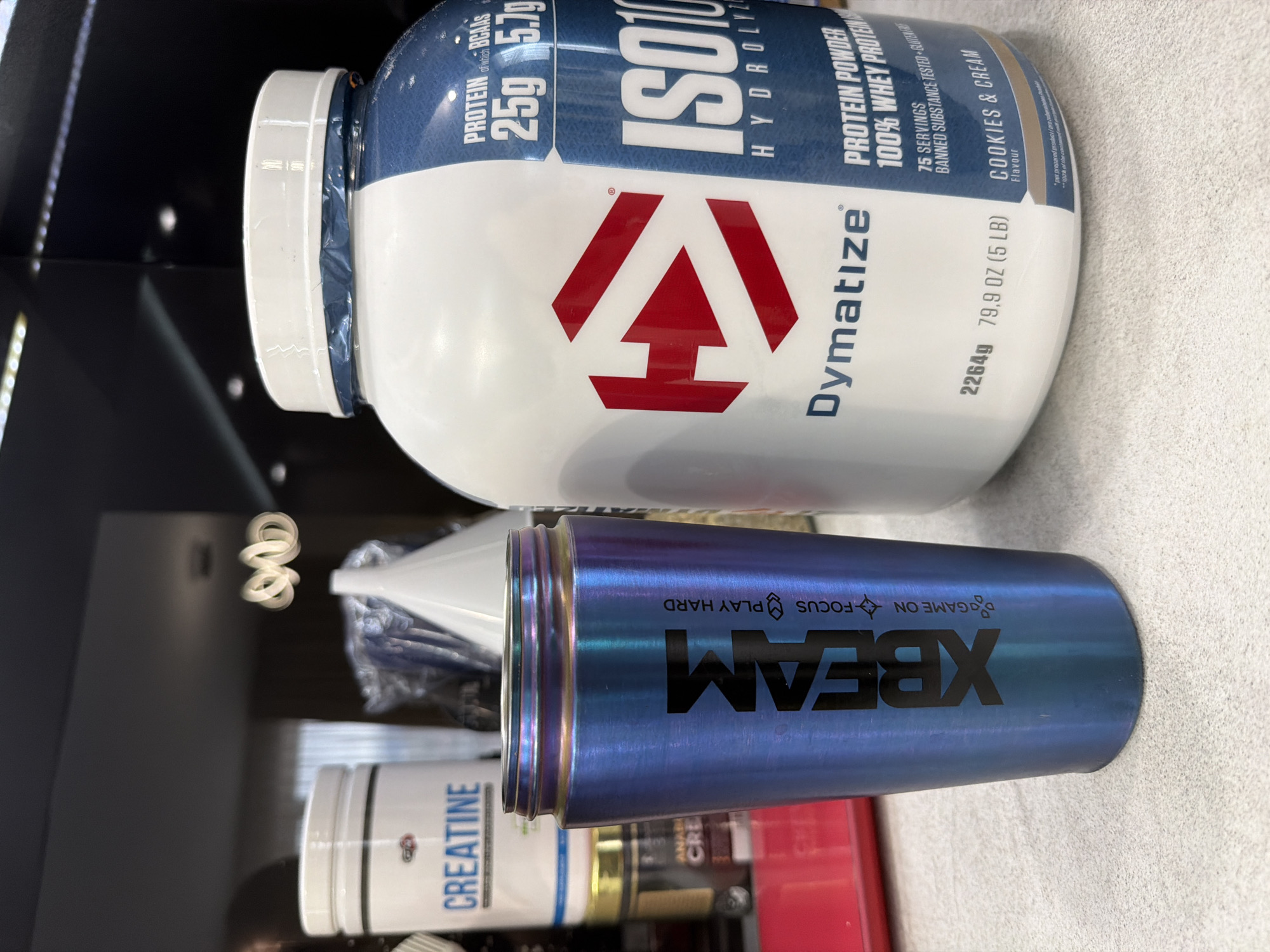 The image shows a collection of fitness supplements and accessories. A large container of Dymatize ISO100 Hydrolyzed Protein Powder in Cookies & Cream flavor dominates the upper part of the image. Below it is a blue metallic shaker bottle with the XBEAM logo printed in black. Partially visible underneath are containers of creatine and other supplements. The scene appears to be indoors, potentially in a cabinet or on a shelf. The setting is impossible to determine.