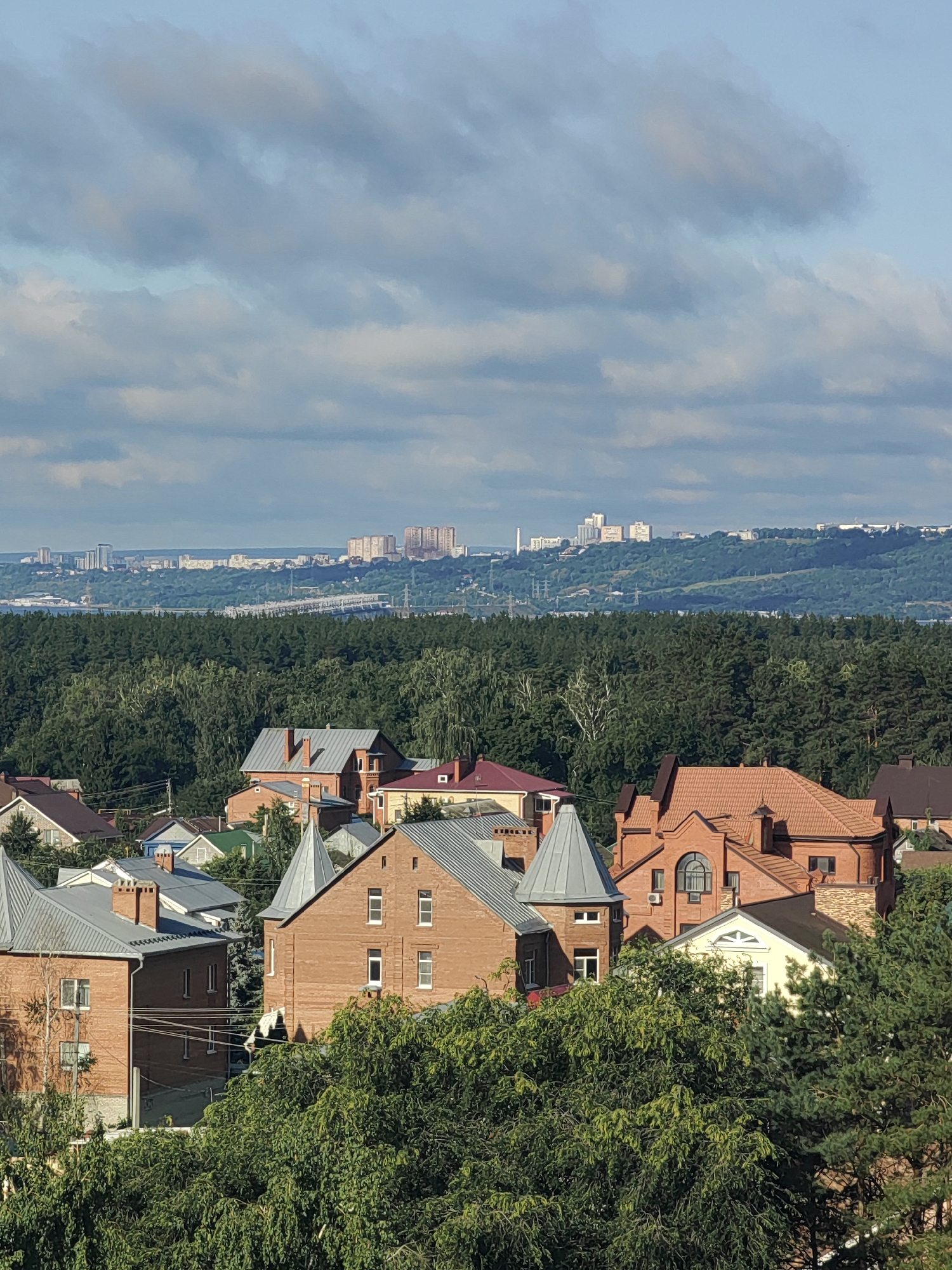 This is an aerial, eye-level, wide shot of a suburban residential area in Ulyanovsk, Russia, with a city skyline visible in the distance. In the foreground, a cluster of red-brick houses with various rooflines, including pointed turrets, are nestled amongst lush green trees. Beyond the houses is a dense forest, and in the far distance, a cityscape with numerous apartment buildings and industrial structures is visible under a partly cloudy sky. The image appears to have been taken on a sunny day.