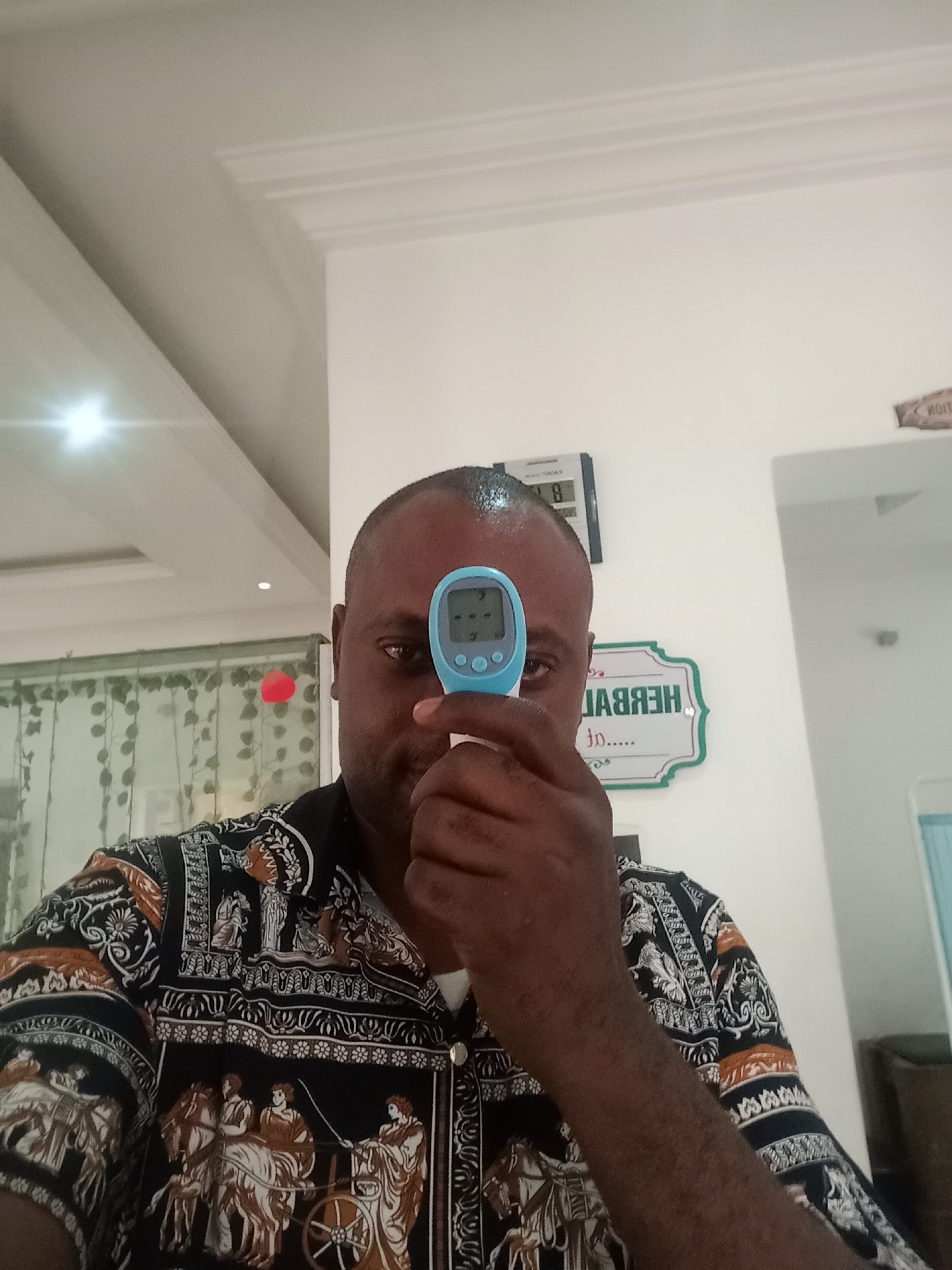 A man stands indoors, holding a blue digital thermometer to his forehead. The thermometer displays a reading. He wears a patterned, collared shirt. Behind him is a white wall. A digital clock is mounted on the wall, showing the time as 8:18. A decorative sign that appears to spell out "HERBAL..." is visible. The lighting is bright, and the environment suggests an interior space. The location is Gwarinpa, Nigeria.