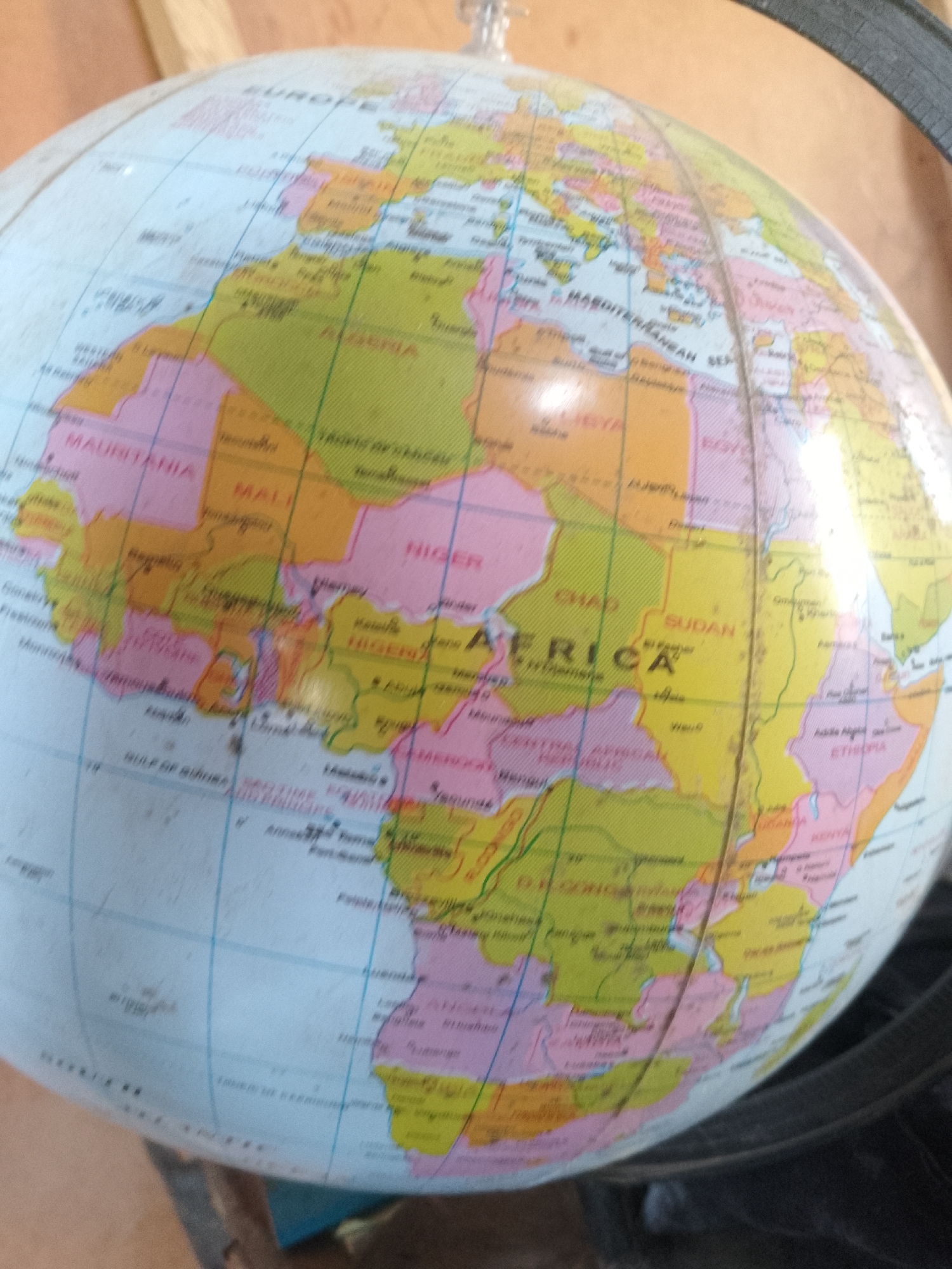 An angled, close-up shot of an antique globe shows the continents of Europe and Africa. The globe is predominantly light blue with colored landmasses divided by black lines. The landmasses are colored yellow, pink, and green, with country borders clearly marked. Text labels are visible in a variety of languages, including "EUROPE," "MAURITANIA," "MALI," "NIGER," "AFRICA," "SUDAN," and "ETHIOPIA." A black stand is partially visible in the lower right corner, and a wooden surface is visible in the upper left. The globe appears to be in a dimly lit indoor environment.