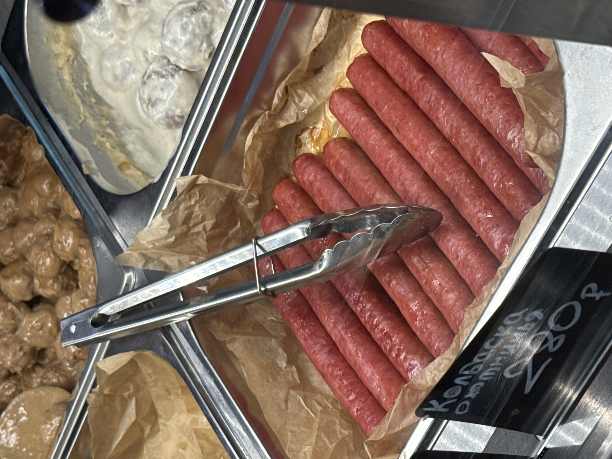 A close-up, overhead shot shows a metal serving tray filled with rows of red sausages, possibly on display in a Yakkima, Russia, food establishment. The sausages are arranged horizontally, filling most of the tray, with some brown parchment paper visible beneath them. A pair of silver tongs rests on top of the sausages, positioned diagonally across the frame. In the background, another tray contains what appear to be meatballs in a creamy sauce. A black price tag in the lower left corner displays text in white, likely a product name and price, though it is partially obscured. The overall scene suggests a self-serve or deli counter in a store or market.