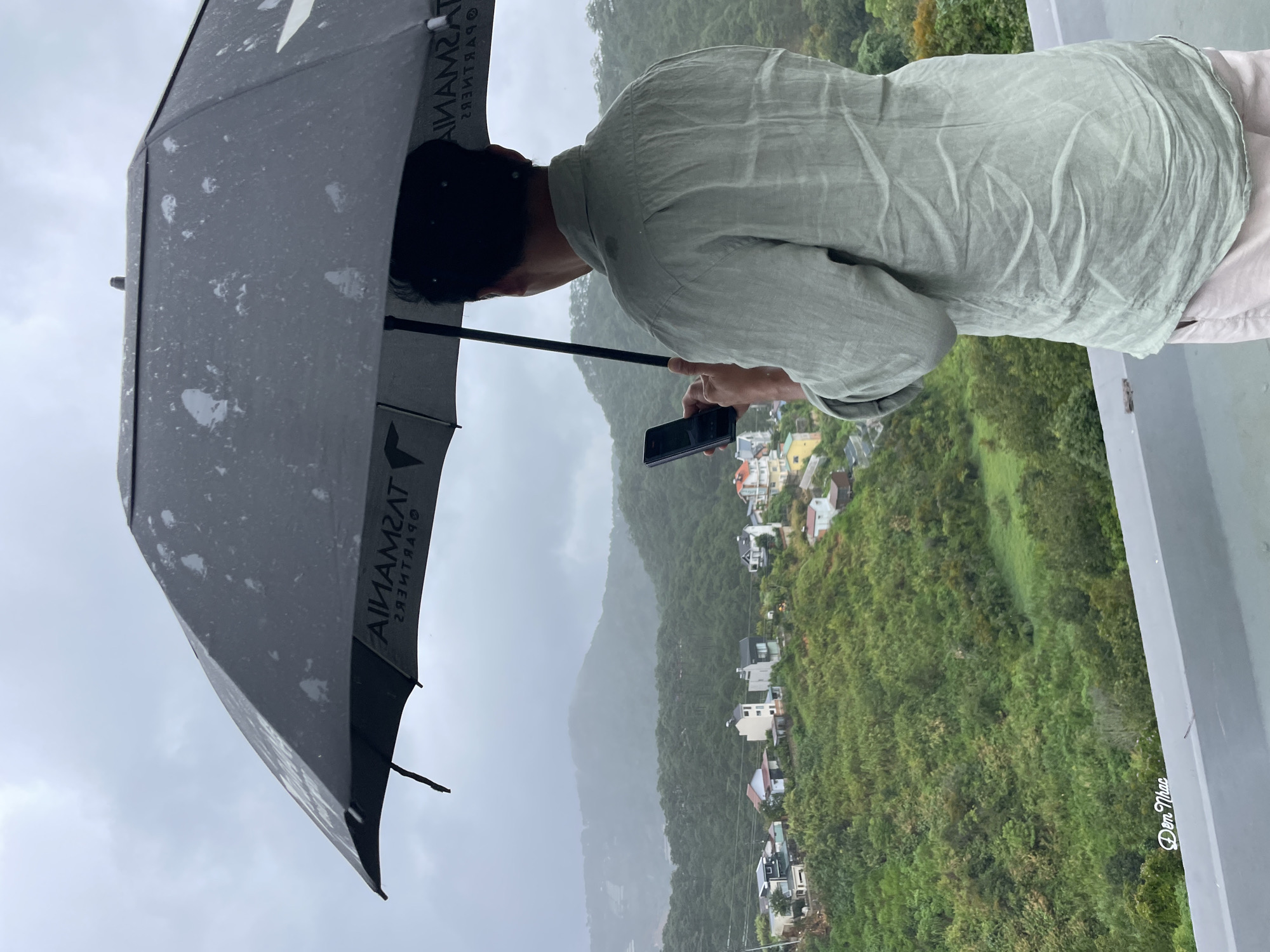 A person stands on a rooftop holding an open umbrella and a mobile phone. The person is wearing a light green linen shirt and light-colored pants. The umbrella, which appears to be black with a logo, is held overhead, protecting the person from the rain. The phone is held in the person's right hand, and the person is looking at the screen.
The background reveals a scenic view of Da Lat, Vietnam. The city is nestled among lush green hills and mountains. Several houses with colorful roofs are scattered across the slopes, indicating a residential area. The weather is overcast and rainy, with visible raindrops on the umbrella. The foreground shows the edge of the rooftop, with a slight shadow visible on the surface. The overall atmosphere is tranquil and slightly melancholic, common in Da Lat during the rainy season.