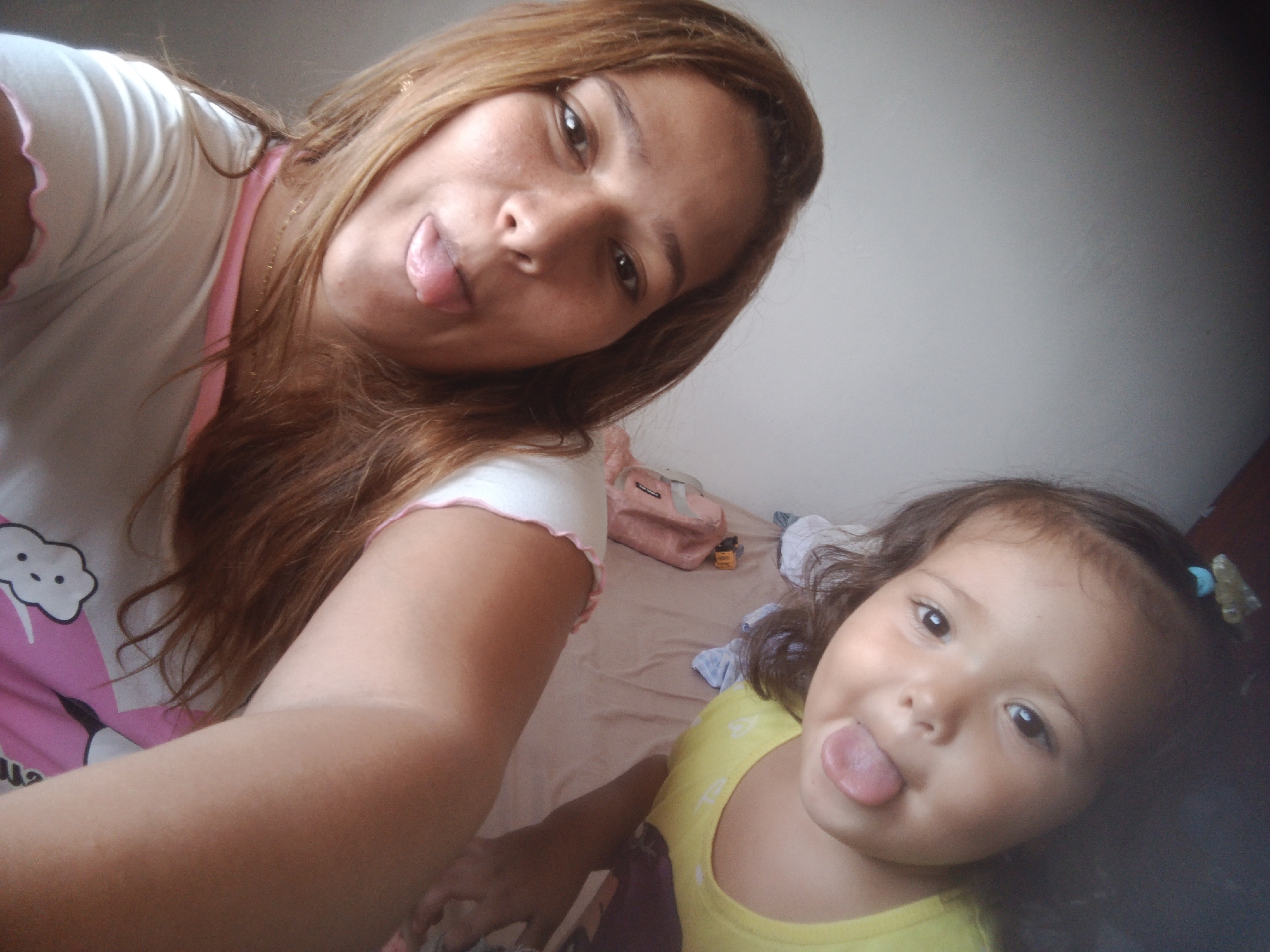 The image shows a selfie taken indoors. Two females are present: an adult and a young child. Both are sticking their tongues out. The adult is positioned on the left, and the child on the right. The background shows a bed covered with a tan-colored sheet, with a pink bag and some scattered clothing items visible. The environment appears to be a bedroom. The location is specified as Caracas, Venezuela.