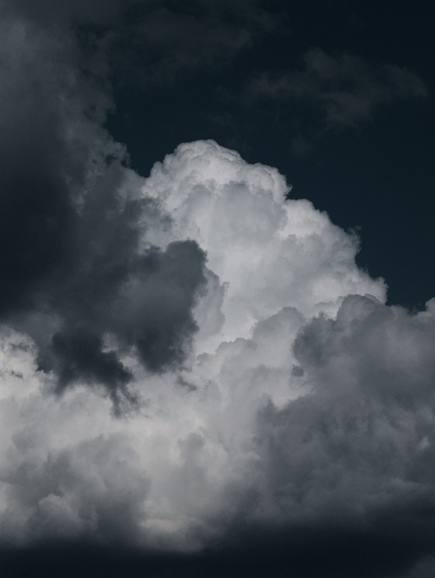 A dramatic, close-up shot captures a dense, tumultuous cloud formation against a dark, twilight sky. The clouds are a mix of bright white, illuminated by an unseen light source, and deep, ominous grays and blacks, suggesting an impending storm. The texture of the clouds is varied, with some appearing soft and wispy, while others are more solid and imposing. The overall impression is one of raw, natural power and the intensity of atmospheric change. The scene appears to be an outdoor setting, likely a sky view from an unspecified location within the city of Unknown, Unknown. There are no people, objects, or actions depicted in the image, only the raw beauty and drama of the natural elements.