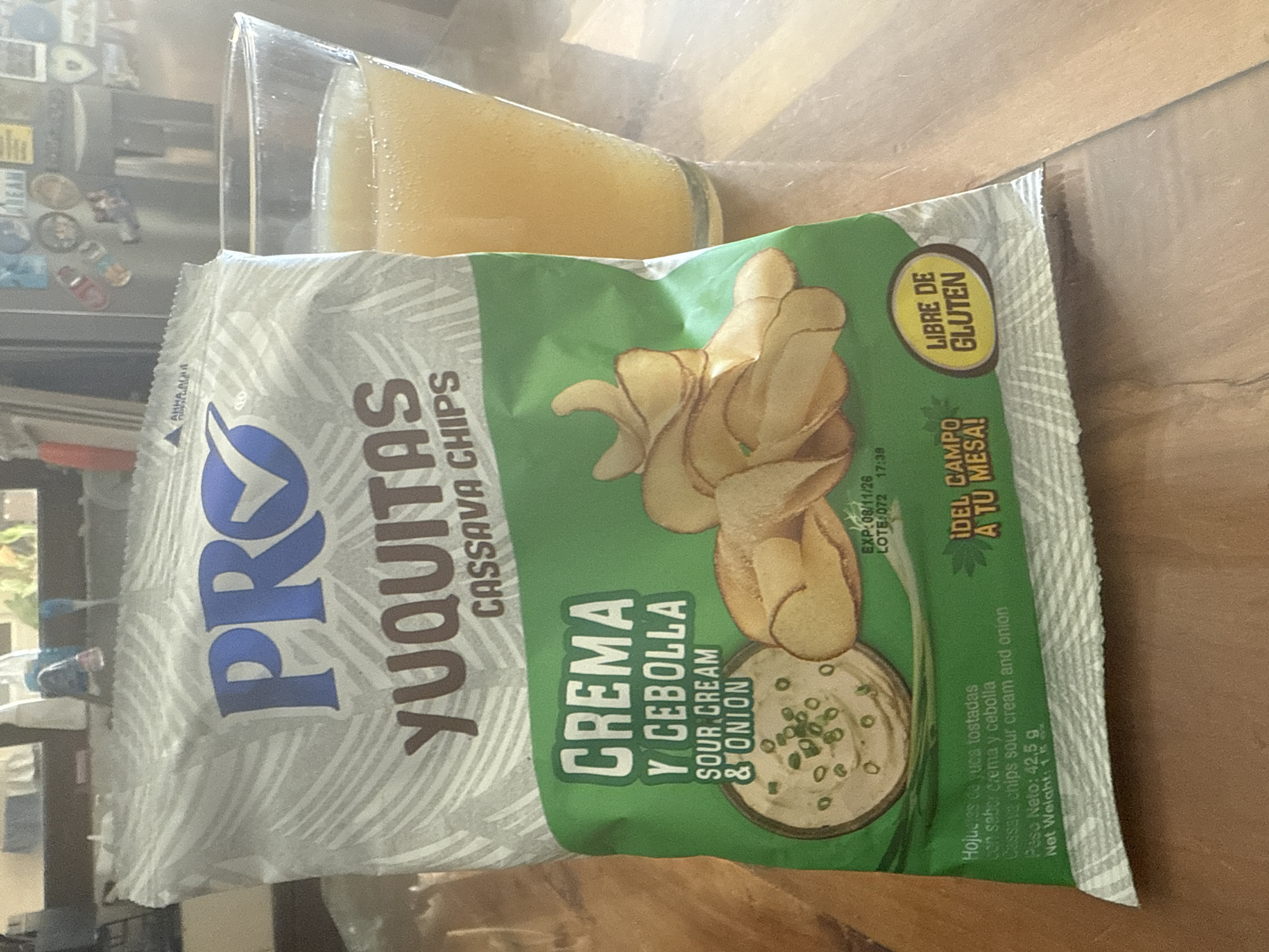 A close-up shot depicts a bag of "Pro Yuquitas" cassava chips with sour cream and onion flavor resting on a wooden surface, partially obscuring a glass filled with a yellowish liquid. The chips are advertised as "libre de gluten," and the bag indicates a net weight of 42.5 grams. The expiration date on the bag is "08/11/26."
Given the product's Spanish labeling ("Crema y Cebolla," "Del Campo a Tu Mesa"), it's plausible that the image was taken in a Spanish-speaking region. However, determining the precise location in Xochitepec, Mexico (or any other city) would require additional contextual clues that are not available in the image.