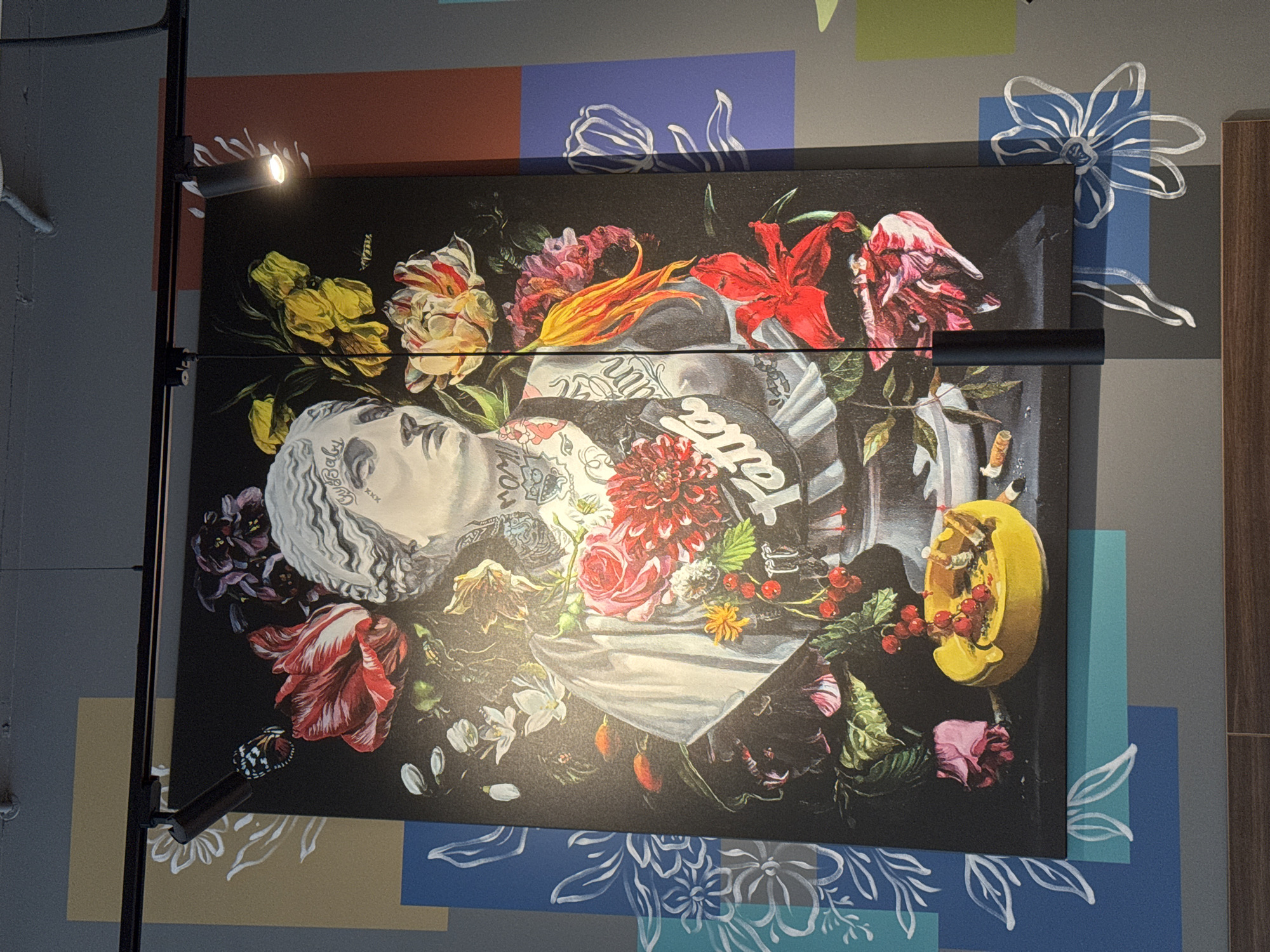The image shows a large painting on a gray wall adorned with a colorful geometric pattern of blue, red, and beige squares. The painting depicts a marble bust of a woman with tattoos, surrounded by an abundance of vibrant flowers and greenery. The woman has light-colored hair pulled back and appears to be wearing a white drape over her shoulders. The tattoos are visible on her neck and chest, with some text and designs. Among the floral elements, there is a butterfly perched on a red tulip, and a bright yellow ashtray with cigarette butts and red berries is situated at the bottom of the painting. Several red and pink flowers, as well as yellow tulips, are prominent in the composition. A black cylindrical light fixture hangs from the ceiling in front of the painting. The wall art is likely located in a commercial establishment in Riga, Latvia, given the context provided.