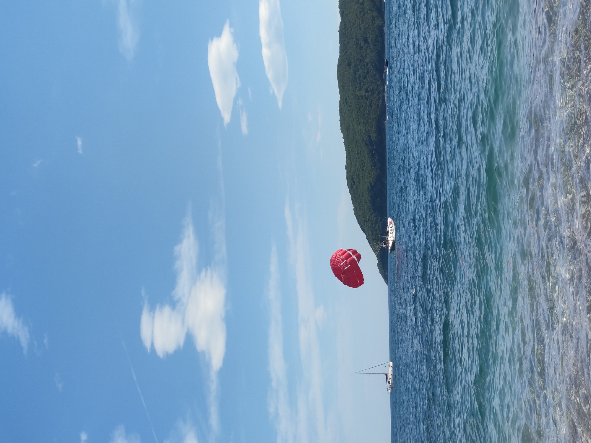 The image depicts a bright, daytime scene at sea in an unknown location. Dominating the frame are the clear blue sky with scattered cumulus clouds on the left and the deep blue sea on the right. A red parasail is floating in the middle of the image, suspended between the sky and the sea, with the suspension cables leading to a boat in the water. A strip of land, likely a forested hillside, runs vertically between the sky and sea. Two boats are visible on the water: a larger, possibly a commercial vessel, is anchored near the hillside, and a smaller sailboat is further out. The sea shows considerable surface texture with light reflecting off the water in areas.