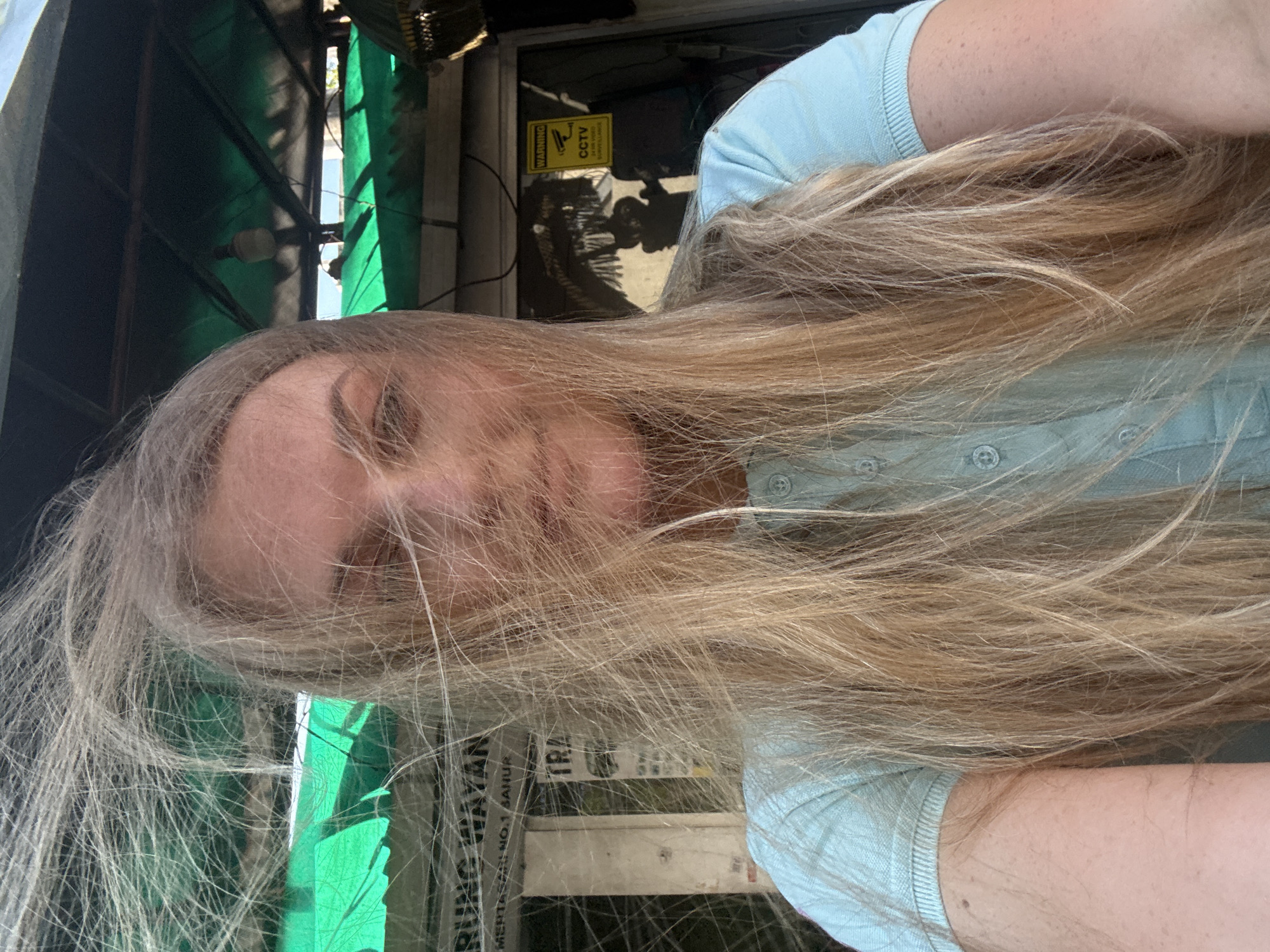 A woman with blonde hair, which is blown by the wind, is taking a selfie outdoors. She is wearing a light blue polo shirt. Behind her, there is a building with a green roof and a sign that reads "RUNG WAYAN MERTASARI NO.1 SANUR". Another sign with a CCTV warning is visible on the right side of the frame. The scene appears to be a street in Denpasar, Indonesia.