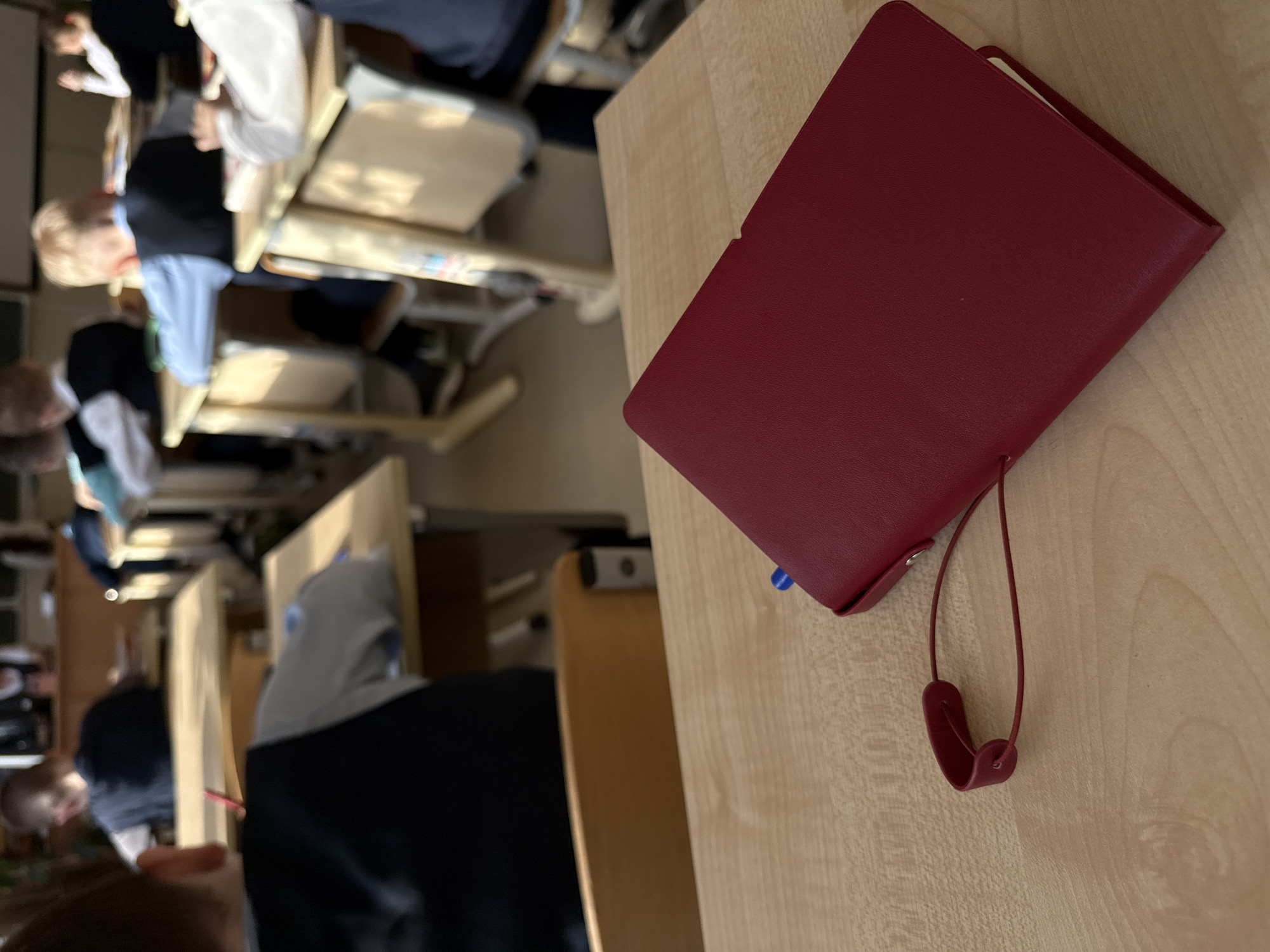 An indoor scene, likely a classroom or study hall, features multiple students seated at wooden desks. The perspective is from a desk in the foreground, looking towards the back of the room.
In the immediate foreground, a distinctive dark red, leather-bound notebook or journal rests on a light wooden desk. A thin, red strap is attached to the notebook, and a blue pen is partially visible, tucked into its side.
Beyond the foreground desk, several rows of similar light wooden desks and chairs are arranged. Numerous students are visible, primarily from the back or side, occupied at their desks. Their attire includes light-colored shirts and darker clothing, suggesting school uniforms or common school wear. The students appear focused, implying an ongoing lesson or study session. The overall lighting is bright and even, typical of an indoor daytime environment. No specific text is discernible on any objects. The scene is set in Perm, Russia.