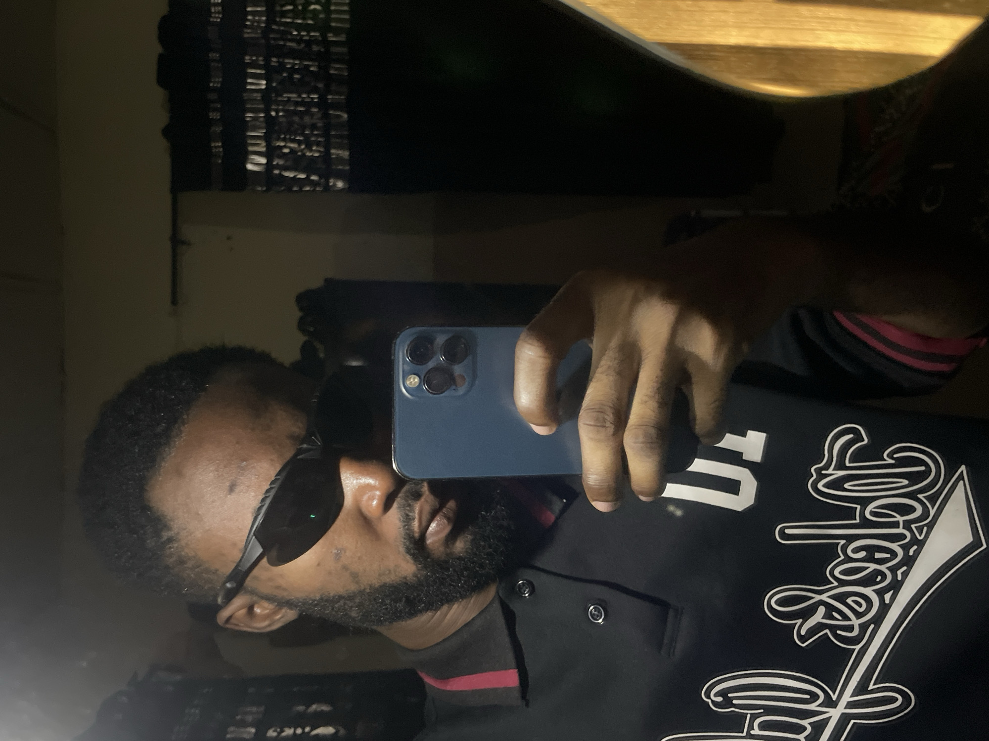 The image depicts a dimly lit indoor scene, likely a bedroom, in Lokoja, Nigeria. The main subject is a Black man taking a mirror selfie. He is wearing a black polo shirt with red and black striped trim on the collar, and sunglasses. He has a beard and appears to be holding a blue smartphone in his right hand to capture the reflection. Behind him, a dark curtain with a shimmering, reflective pattern is visible, and to his right, a portion of a wooden object, possibly furniture, is illuminated. The overall ambiance is dark, suggesting it is nighttime or the room has limited lighting.