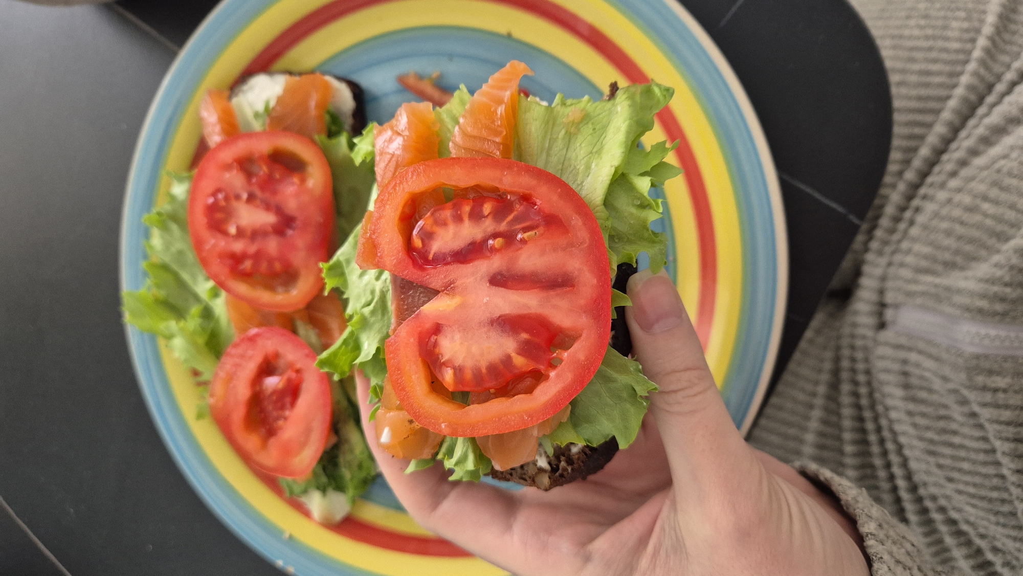 A hand holds a sandwich of rye bread, lettuce, salmon, and a tomato slice. The sandwich is on a plate with additional sandwiches of similar construction. The plate is multi-colored with bands of red, yellow, and blue. The scene includes a dark-colored table and a person wearing a grey sweater. The location is not identifiable as Batumi, Georgia, based on the visual information provided.