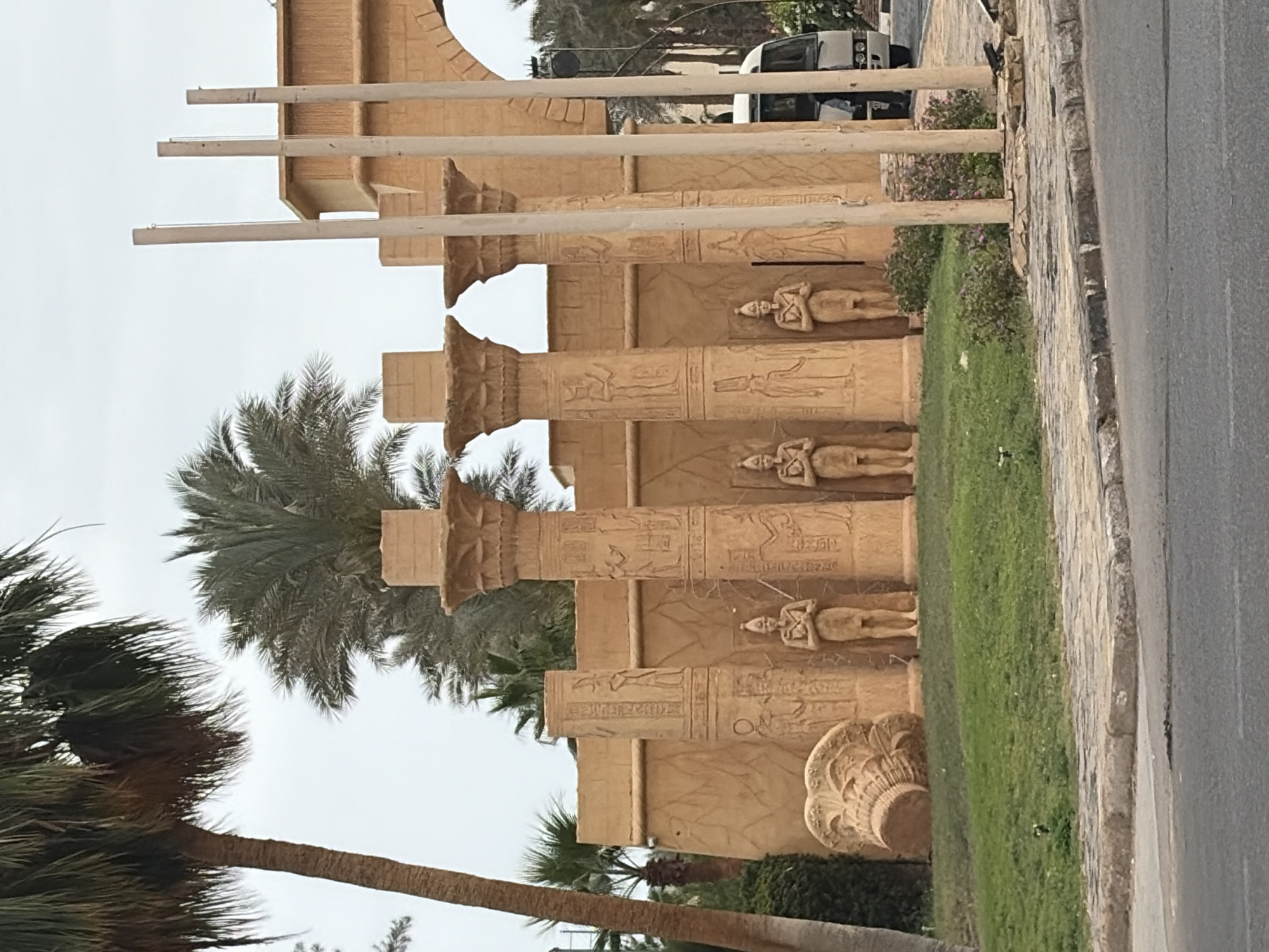 The image shows a section of architecture in Sharm El Sheikh, Egypt, adorned with pharaonic-style designs and hieroglyphs. Three pillars with detailed capitals stand prominently, each decorated with carved figures. Lush palm trees frame the architectural structure, contrasting with the beige tones of the building. A manicured lawn and a section of roadway abut the architecture.
