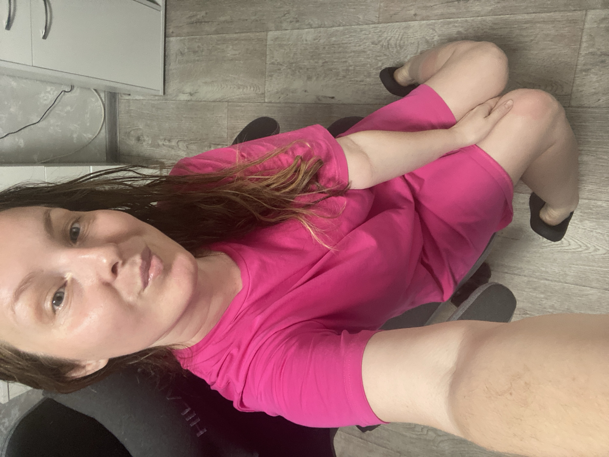 A woman is seated on a chair in an indoor setting. She is wearing a bright pink shirt and dark shoes. The flooring is light grey wood-patterned material. In the background is a white cabinet with silver handles. The location is Noviy, Russia.