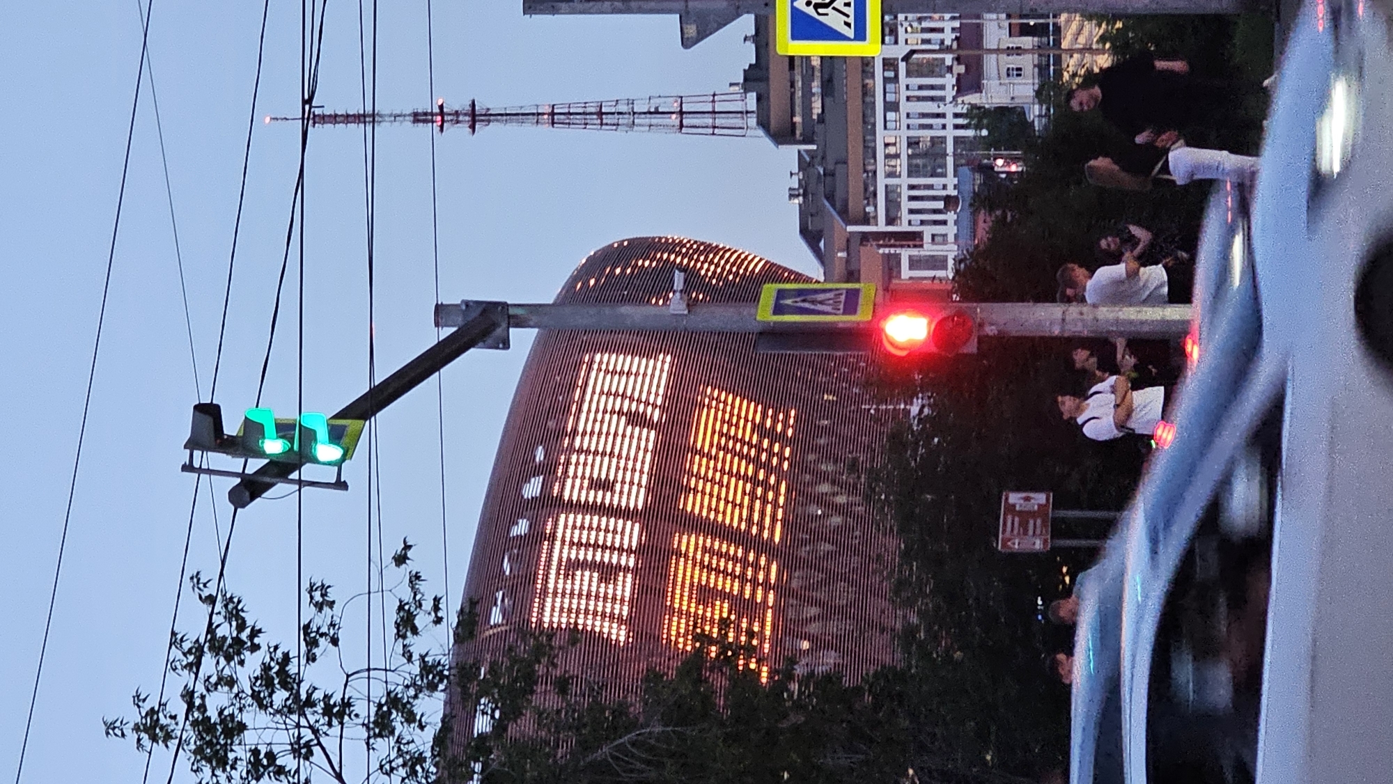 The image captures an evening street scene in Yekaterinburg, Russia, dominated by a modern, curved building illuminated with bright, pixelated text that appears to read "ЛЕТО ПАРК" (Summer Park) in Cyrillic script. A traffic light in the foreground is showing red, indicating a stop. Several people are gathered near the traffic light, some standing and talking, others waiting. A white car is in the foreground, partially blurred due to motion. In the background, a tall radio tower with red lights is visible against a twilight sky. The overall environment suggests a public space in a city setting.