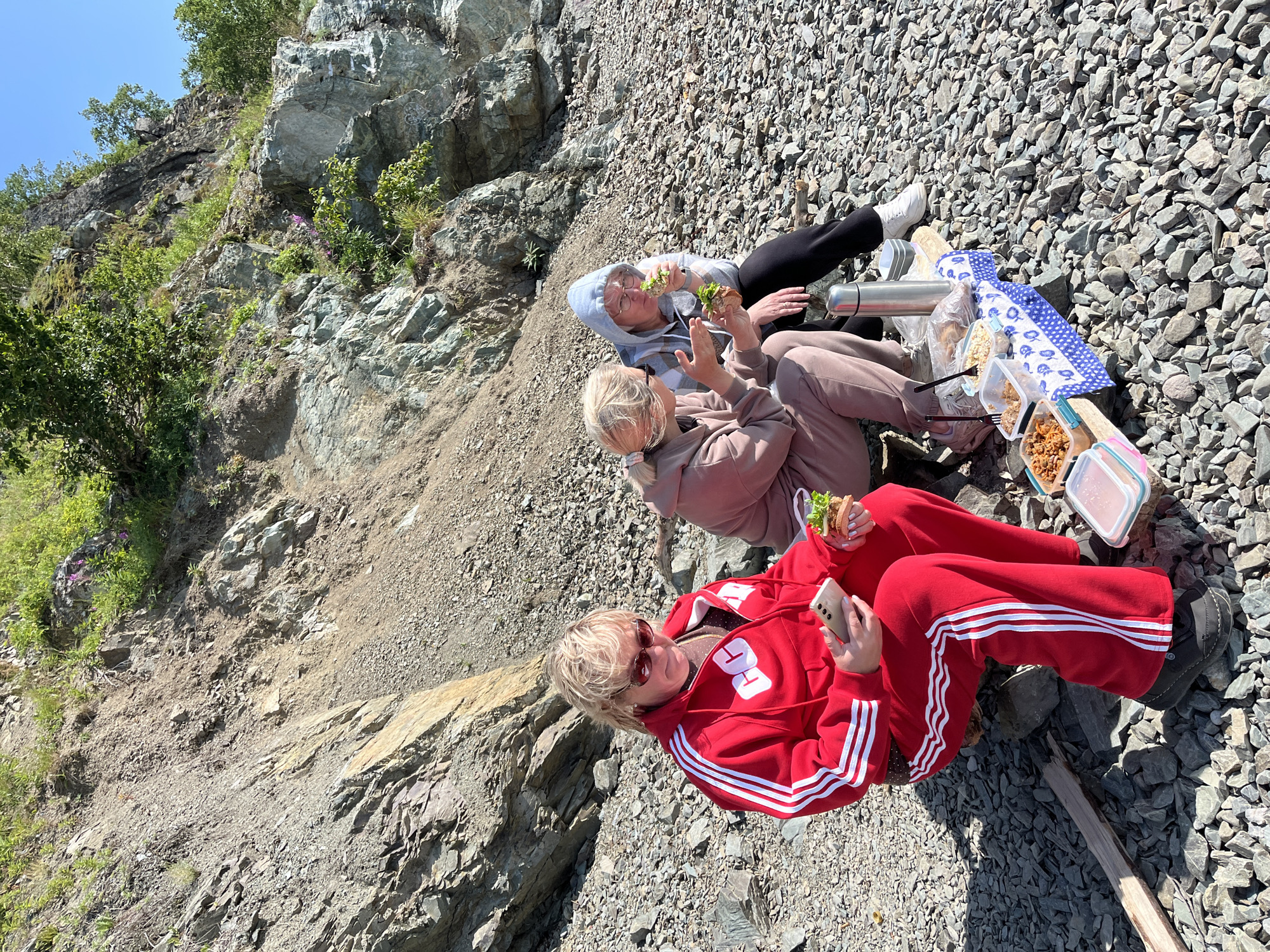 Three women are having a picnic on a rocky shore, with a steep, overgrown hillside behind them. The woman in the foreground wears a red tracksuit and sunglasses, and is holding a smartphone and a sandwich. The woman to her left wears a light brown tracksuit and is also holding a sandwich. The woman on the far right wears a grey hooded sweatshirt and black pants, and is eating a sandwich. A blue and white checkered tablecloth is spread out on a wooden plank, holding various food containers and a thermos. The rocky terrain suggests a natural, potentially coastal or mountainous environment. This scene is consistent with outdoor recreational activities observed in the outskirts of Petropavlovsk-Kamchatsky, Russia, given the geographical features and the type of clothing worn, which often reflects practical choices for cooler climates and outdoor excursions.