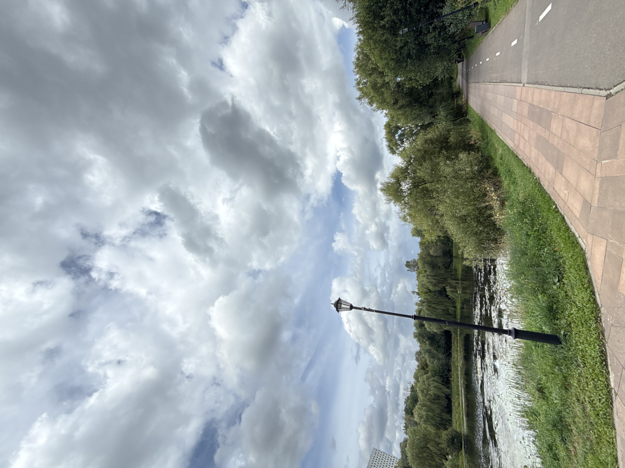 This image depicts a park scene in Moscow, Russia, likely taken on a cloudy day. In the foreground, a paved path curves through the park, made of reddish-brown square tiles on the left and asphalt with white lane markings on the right, suggesting a dedicated bicycle or running path. A dark lamppost stands to the left of the path. To the left of the path is a body of water, possibly a pond or lake, with lily pads and aquatic vegetation. A dense line of green trees and foliage borders the water. In the background, more trees and a tall, modern building with a grid-like pattern are visible on the far left. The sky is filled with dramatic, heavy clouds, indicating overcast conditions. There are no people or animals visible in the frame.