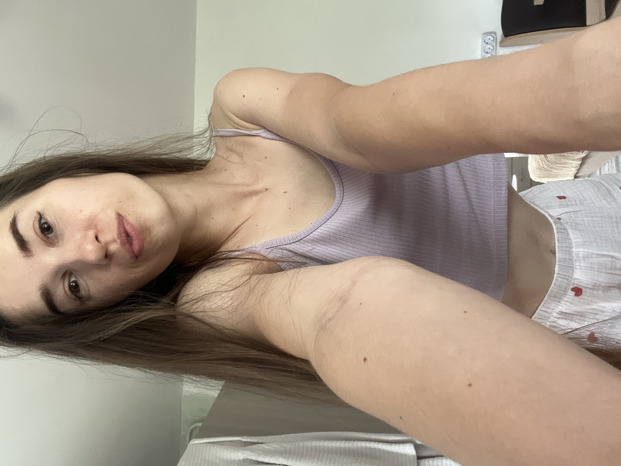 A young woman with long brown hair, wearing a light purple tank top and white shorts with small red hearts, is taking a selfie in what appears to be a bedroom. She is positioned close to the camera, with her arms extending outwards, one closer to the lens than the other. The background shows a light-colored wall, a closet with clothing hanging on the left, and a power outlet on the right. There is a light-colored headboard or furniture visible in the background. The lighting is soft and natural, suggesting an indoor environment during the day.