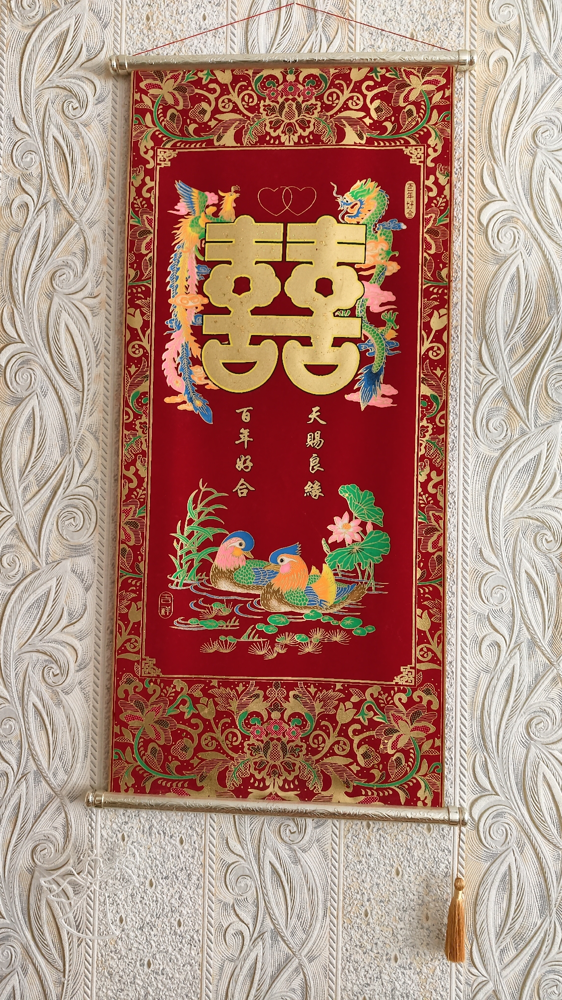 A red scroll with gold and colorful Chinese motifs hangs on a textured white wall. The scroll features a large gold "double happiness" character with two intertwined hearts above it. Flanking the character are a dragon on the right and a phoenix on the left. Below the character, two Mandarin ducks are depicted in water with lotus flowers and reeds. To the left of the ducks, two vertical lines of Chinese calligraphy read "百 年 好 合" (bǎi nián hǎo hé), meaning "a hundred years of harmony." To the right, two vertical lines read "天 賜 良 緣" (tiān cì liáng yuán), meaning "heaven-granted good match." The scroll has gold decorative borders and a gold tassel hangs from the bottom right corner. The wall has a raised relief pattern of stylized leaves and vines. The scene is illuminated by bright, even lighting, casting no discernible shadows. The image does not contain any people or provide any information about its location in Horishni Plavni, Ukraine.
