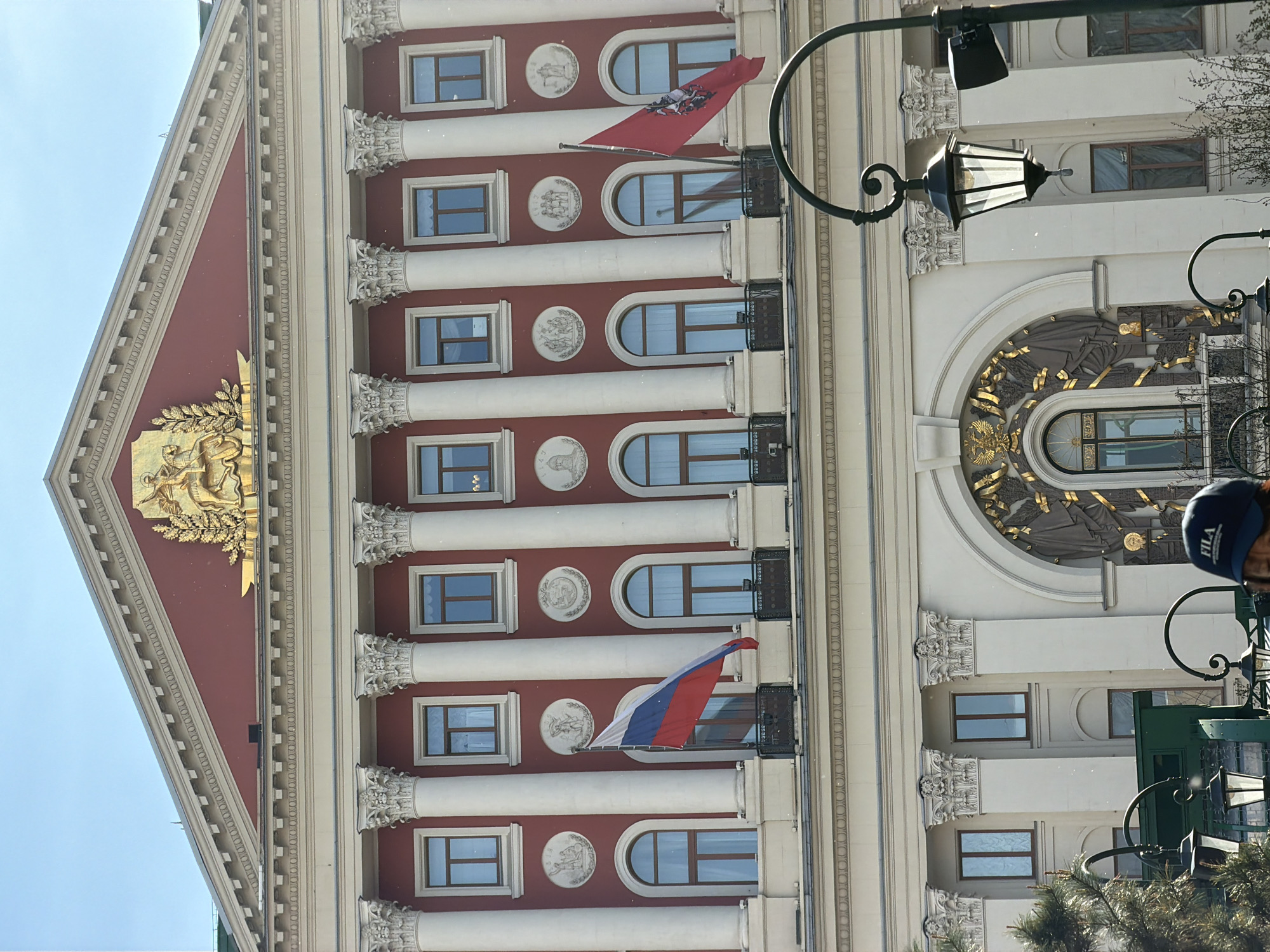 The image depicts a fragment of the Moscow City Duma building in Moscow, Russia. The building facade is characterized by a red and white color scheme, featuring Corinthian columns and decorative moldings. The building displays windows and medallions and is adorned with the flags of Moscow and Russia. A vintage street lamp is visible in the foreground. A person's head can be seen in the foreground as well. The sky is clear.