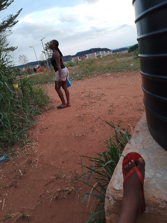 The image shows an outdoor scene in Ogboko, Nigeria. In the background, a woman wearing a dark top and light shorts stands on a red dirt path, looking upward toward a tree. She has a blue object in her hand. Behind her are buildings. In the foreground is a water tank and a person's leg with a red flip-flop. The ground is red dirt, and there is some grass and other vegetation present. The sky is overcast.