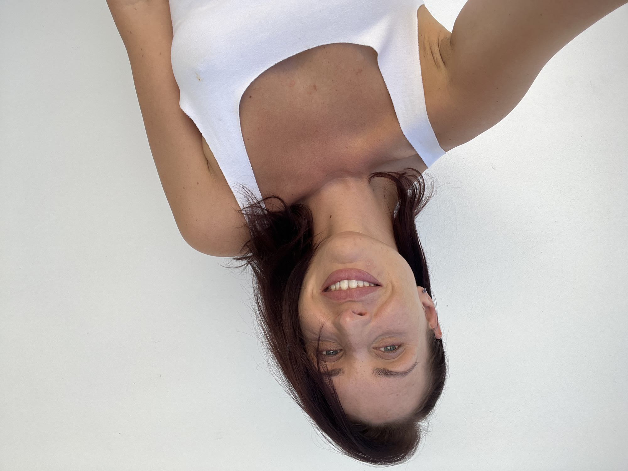 A woman with dark hair, light-colored eyes, and a tanned complexion is depicted in a brightly lit, headshot-style portrait. She is smiling broadly, revealing her teeth, and appears to be looking directly into the camera. Her dark hair falls forward on the left side of her face, partially obscuring her left eye. She is wearing a simple white tank top or camisole, which contrasts with her skin tone.
Her head is tilted slightly, and her upper body is framed against a plain, stark white background. Her arms are raised and visible on either side of her, extending upwards from the bottom of the frame. Notable details include some faint reddish marks on her chest and a small, light brown stain on her white top near her right armpit. The overall impression is one of a cheerful and friendly individual captured in a straightforward, well-lit photograph. There is no visible text in the image. The setting appears to be minimalist, possibly indoors or against a very clean white surface.