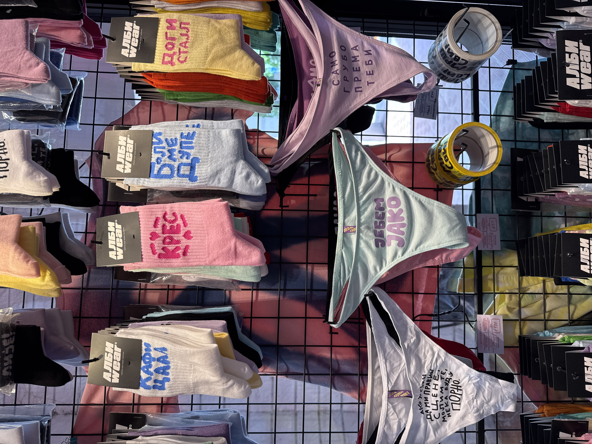 The image shows a display of socks and underwear hanging on a black wire grid in what appears to be a shop setting. The socks, packaged with "АЛБИ WEAR" tags, come in various colors and have text printed on them. The underwear pieces, also displayed on hangers, have messages written on them in what appears to be Serbian. Two rolls of tape are also on display, with Serbian text printed on the tape. The scene suggests the location could be a retail space in Novi Sad, Serbia, based on the Cyrillic script used in the text on the items. The background appears to be a blurred image of people.