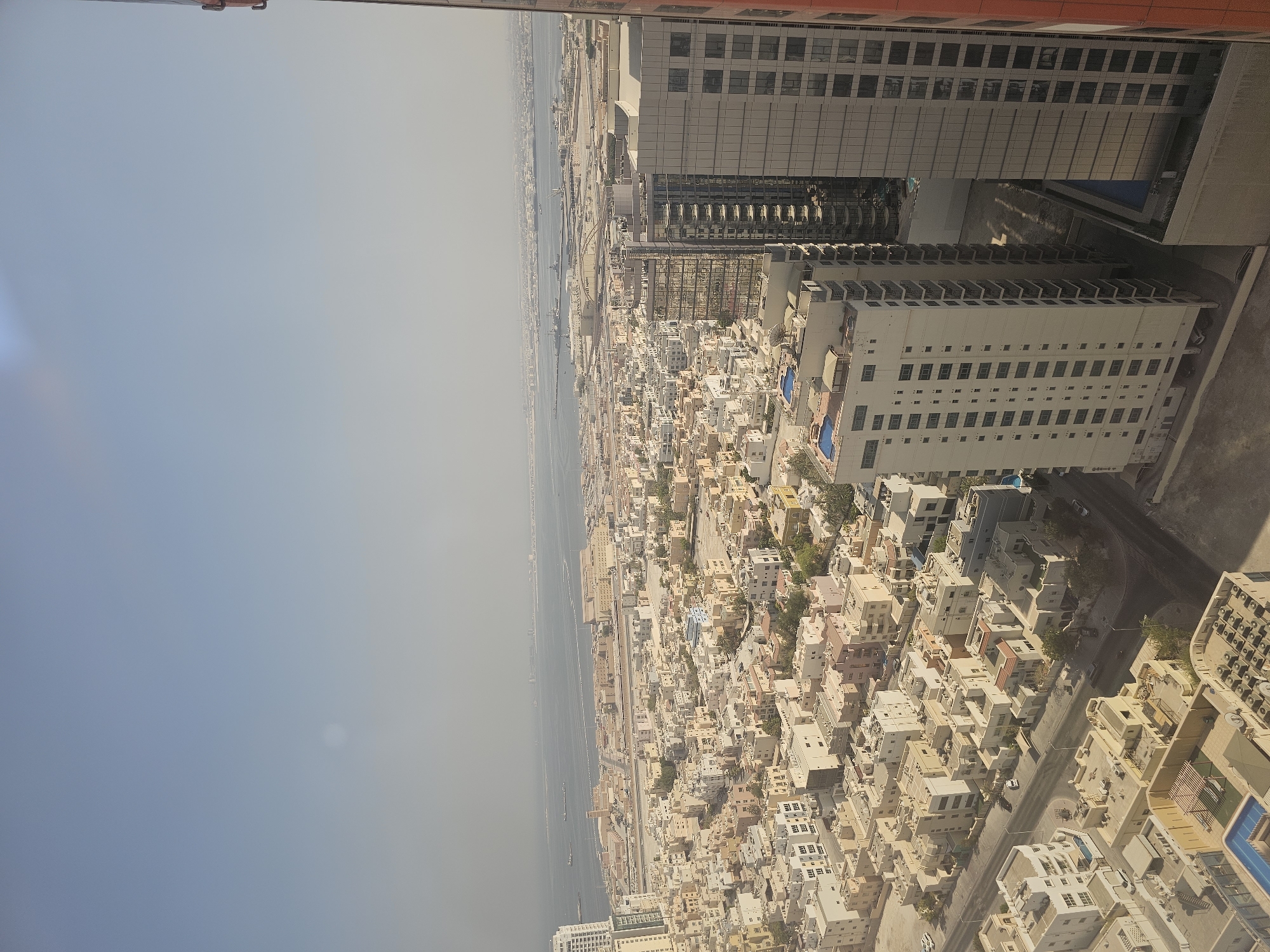 An aerial, wide-angle photograph showcases a dense urban landscape in Manama, Bahrain, with a hazy sky overhead. The foreground features modern, light-colored residential buildings with numerous windows, some with balconies. One prominent tall building on the right has a reflective, dark glass facade. Below these skyscrapers, a sprawling neighborhood of lower-rise, beige and white buildings with flat roofs and many windows is visible. Streets with sparse car traffic wind through this area. Several buildings have rooftop swimming pools. In the mid-ground, a more extensive complex of lighter-colored, uniform buildings stretches towards the waterfront. Beyond this, a large body of water, likely the Arabian Gulf, is visible, with a distant cityscape and industrial structures on the horizon. The overall impression is one of a developed, coastal city under a hazy, possibly polluted, atmosphere.