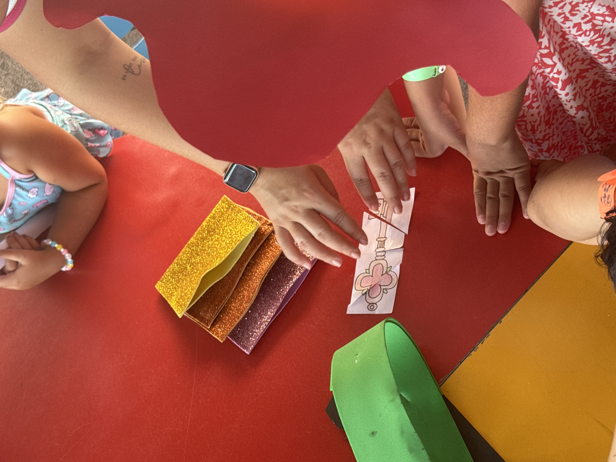 The image shows a close-up view of hands of various individuals, presumably children and adults, engaged in a craft activity on a red table. Glitter paper in shades of yellow, orange, and purple is stacked in the upper right quadrant of the image. A small, printed image of a key with a flower design is in the center, near the hands. To the left, a green, curved object, possibly a piece of craft material or a container, is visible. The overall scene suggests a creative or educational setting. The location of Aksu, Turkey, is not determinable from the visual information provided in the image.