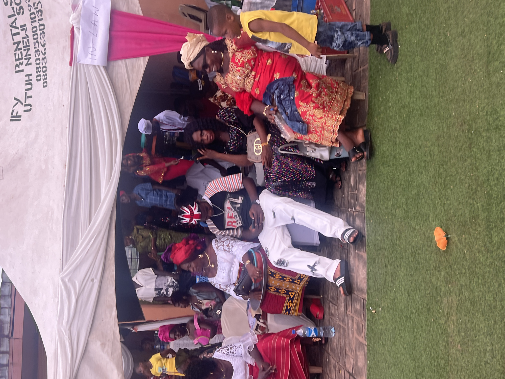 The image captures a gathering of people, seemingly at an event labeled "In-Law", as indicated by a sign. The crowd is under a tent structure with white and pink draping. Several individuals are seated, while others are standing. Some people are dressed in traditional attire. The environment appears to be a paved outdoor area, adjacent to a patch of artificial turf. Contact information for IFY RENTAL SC UTUH NNEWI is visible on the side of the tent. The precise location within the city of Unknown, Unknown, cannot be determined from the image.