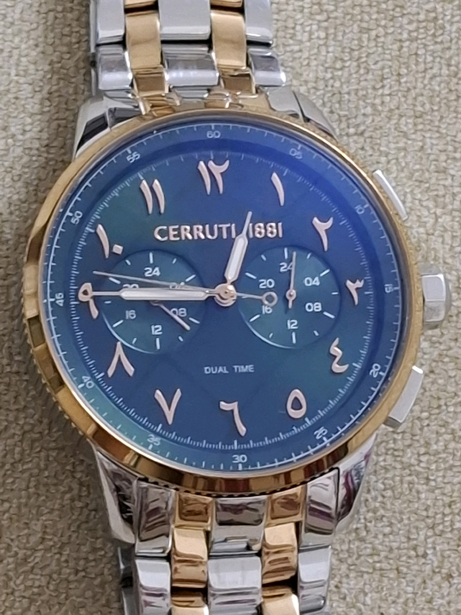 A close-up, eye-level, portrait-oriented photograph shows a wristwatch with a blue face and gold and silver band. The watch has Arabic numerals. There are three subdials on the watch face, with the numbers 24, 16, 12, 04, and 08 visible. The brand name "CERRUTI 1881" is visible across the center of the watch face. The watch is resting on a textured, beige surface. There is no indication of the city of Abu Dhabi, United Arab Emirates, or any other location information present in the image.