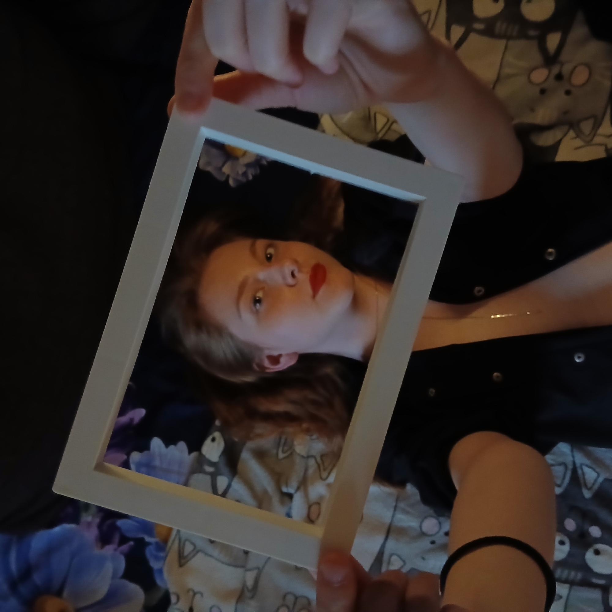 An angled, close-up indoor shot features a person, likely a young female, looking upwards and directly at the viewer. Her face is framed by a white rectangular border held by her hands. She has fair skin, light brown hair, and is wearing red lipstick, with her eyes wide open.
Her hands grasp the top and bottom edges of the white frame. She is dressed in a dark, possibly black, garment adorned with visible metal studs or buttons. A delicate, thin necklace is visible around her neck. A black hair tie is worn on her right wrist, which is seen in the lower right portion of the image.
The background inside and outside the frame consists of a patterned fabric, likely bedding or a blanket, featuring cartoonish white and beige cat faces with prominent eyes. In the lower-left corner, outside the frame, a soft, purple-blue textured object, possibly a decorative pillow or stuffed animal, is partially visible. The lighting suggests an indoor setting, possibly with soft ambient or artificial light. There is no visible text. The overall scene depicts a casual portrait.
