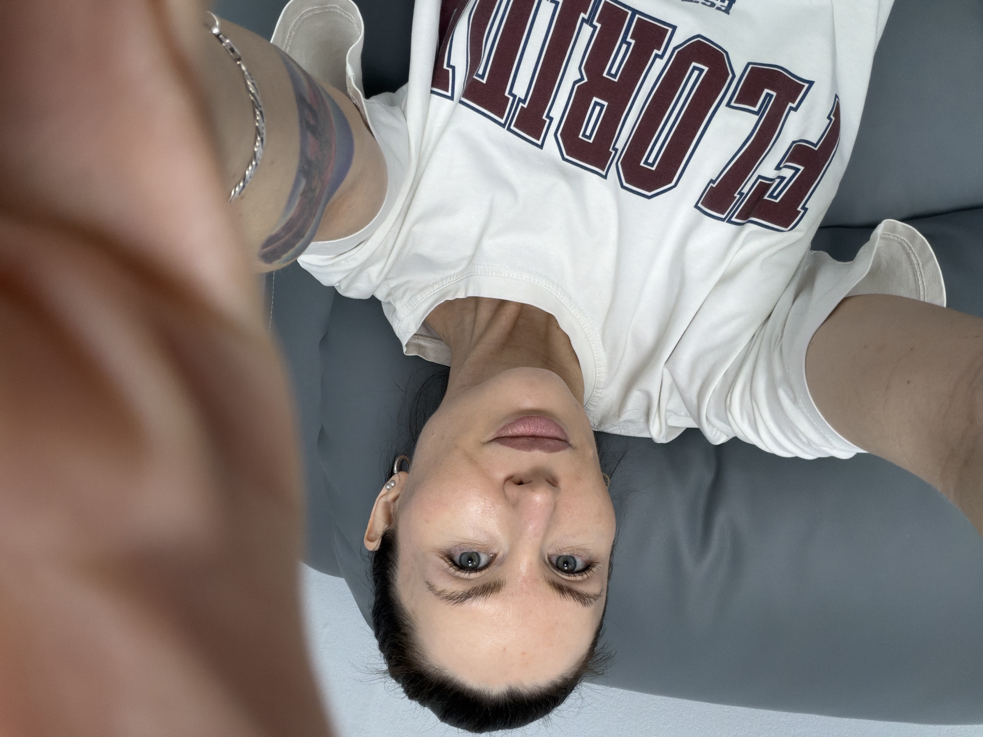 A woman with fair skin, dark hair pulled back, and blue eyes is lying on a gray surface, possibly a sofa or bed. She is wearing a white t-shirt with the word "FLORIDA" in maroon lettering with a blue border. She appears to be taking a selfie, as a blurred hand and arm are visible in the foreground. A silver bracelet is around her left wrist, along with a partial view of a tattoo on her upper left arm. A small pearl earring is visible on the right side of her face. The location cannot be determined from this image, so I cannot include Perm, Russia.