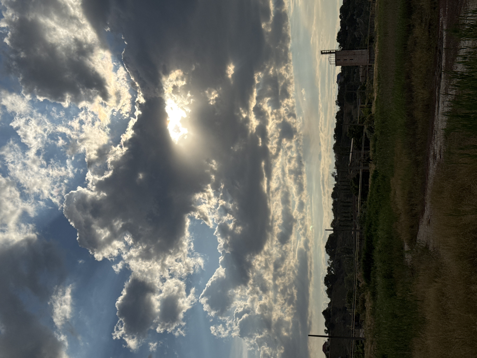 This media file depicts an outdoor scene with a dramatic sky dominating the frame. The sky is filled with a mix of dark, brooding clouds and brighter, sunlit clouds, suggesting a dynamic weather pattern. Rays of sunlight break through the clouds, creating a luminous effect.
The lower portion of the image shows a landscape that appears to be either rural or undeveloped. There is a grassy field, some of which appears dry and cracked, possibly indicating a dry spell or a parched area. In the distance, a structure resembling a small tower or silo is visible, with a simple architectural design. Trees and a hillside form the backdrop to this structure. There are also some industrial-looking elements such as metal poles and possibly remnants of fencing or tracks.
The scene suggests it is either late afternoon or early evening, given the quality of the light and the cloud formations. The overall mood is somewhat dramatic due to the interplay of light and shadow in the sky. The location is identified as Odesa, Ukraine. No people are visible in the image, and there is no visible text.