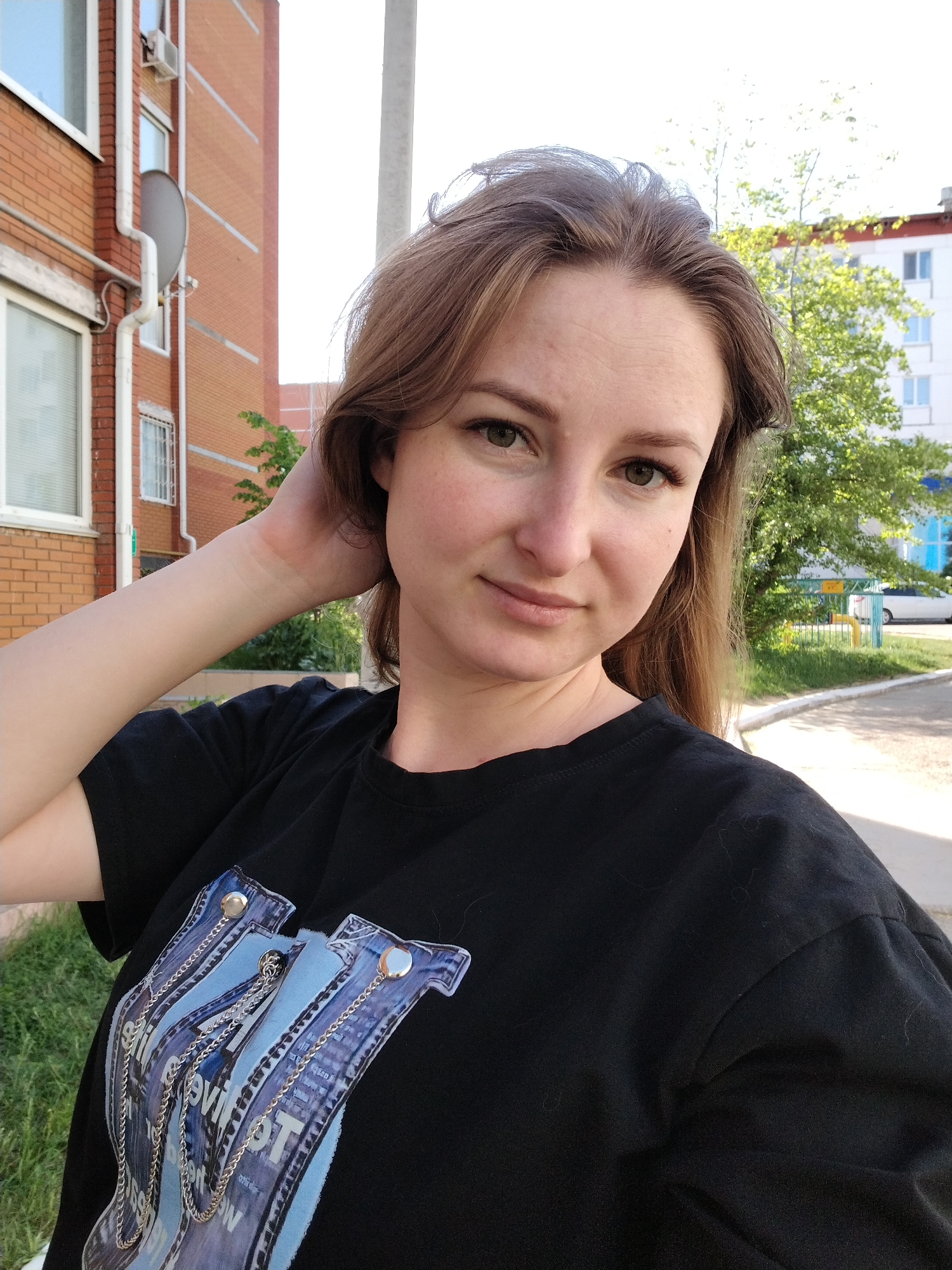 A young woman is the focus of an eye-level outdoor shot. She is fair-skinned with brown hair and is wearing a black T-shirt. Her right hand is raised, touching the back of her head, and she is gazing directly at the camera with a slight smile.
Behind her, a typical urban scene unfolds. A brick building with windows and a satellite dish is visible on the left. A tall utility pole stands in the background. A leafy tree and a white building can be seen to her right. The scene is well-lit, suggesting daytime, and the location is potentially within the city of Chaikovsky, Russia.