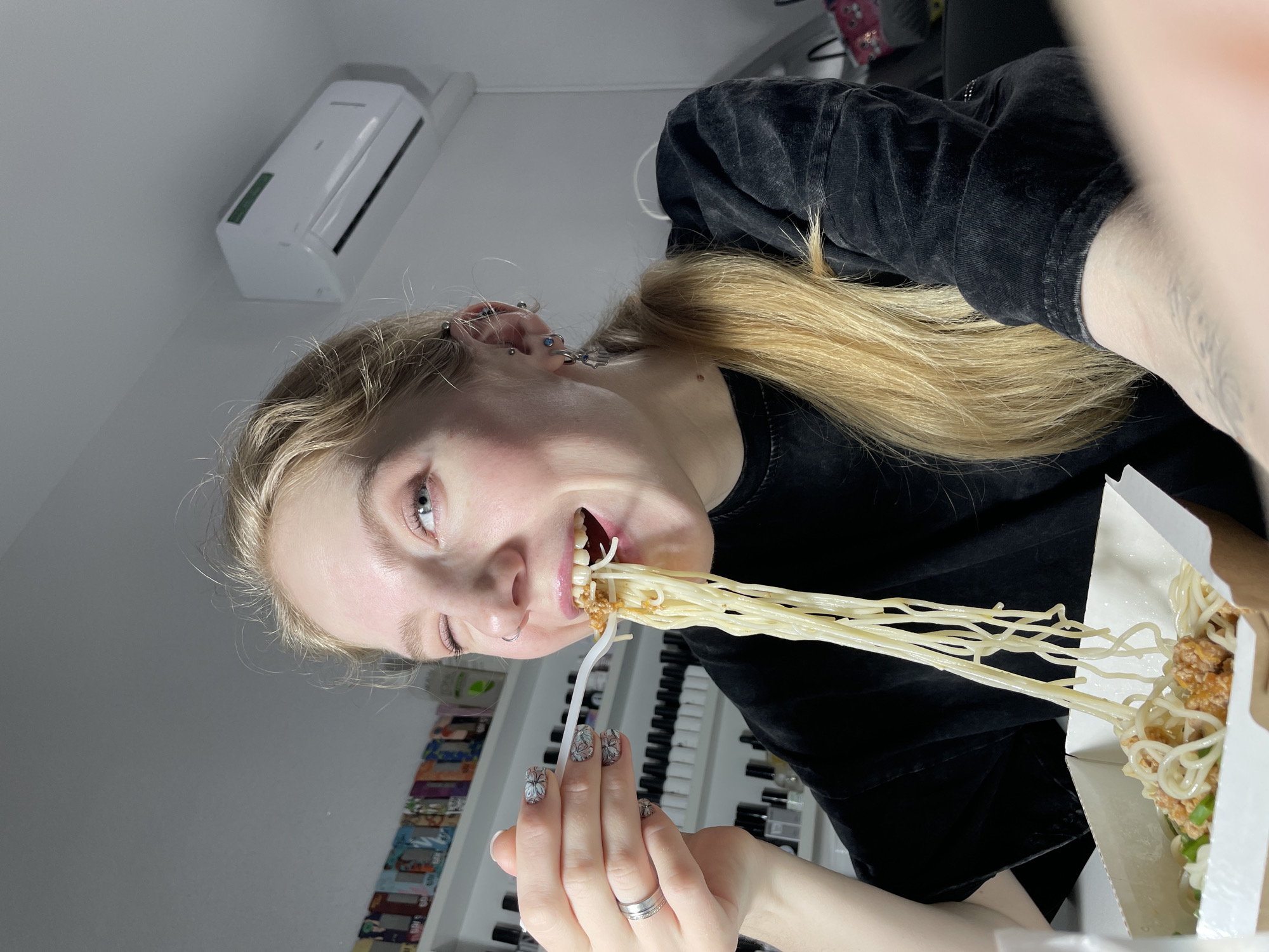 A young woman with light skin and blonde hair pulled back in a low ponytail is in the midst of eating a forkful of long, thin noodles. She has multiple ear piercings and a ring on her finger. The noodles are coming from a white takeout container, which also seems to contain fried chicken and some green garnish. The woman is wearing a black top. In the background, there is a wall shelf with what appears to be an assortment of nail polish bottles or similar products. There is also a white air conditioning unit mounted on the wall. The location, Izhevsk, Russia, cannot be determined from the visual information.
