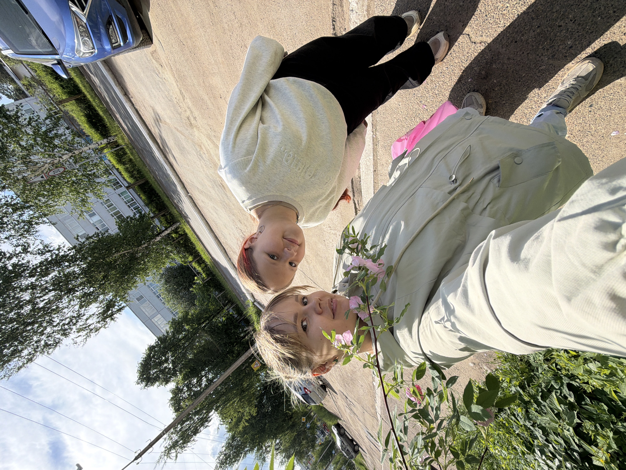 Two individuals are seen in a selfie-style photograph taken outdoors on a street in what is likely Chaikovsky, Russia. One individual is wearing a light green jacket and light blue jeans. The other is wearing a light gray sweatshirt and black pants. They are standing next to a bush with pink flowers. A street, buildings, and parked cars are visible in the background. The photograph appears to be rotated counterclockwise.