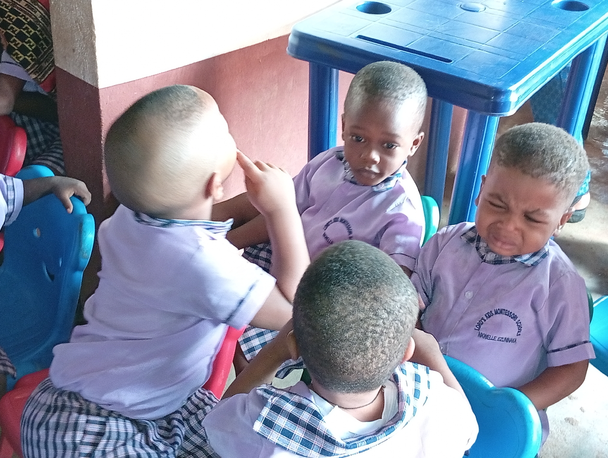 Four young children are seated around a small, blue, plastic table, likely in a classroom setting in Unknown, Unknown. The children are all wearing light purple short-sleeved shirts with checkered collars, possibly a school uniform, bearing the name "LORD'S KIDS MONTESSORI SCHOOLS, NKWELLE EZUNAKA." One child has his back to the viewer. Another has tears and a scrunched face. A third is looking at the first with a neutral expression. The fourth is touching the first child's face with his index finger. The children are seated on small, brightly colored plastic chairs. A wall with a maroon section is visible in the background.
