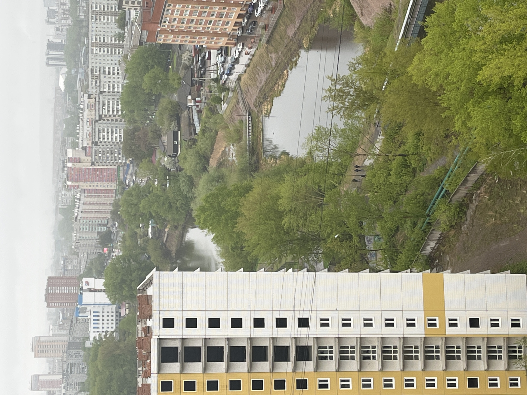 The image depicts an urban scene in Unknown, Unknown. In the foreground, a tall building is visible, with sections of white and yellow panels. Many windows are present, some filled with dark gray concrete. Adjacent to the building is dense green vegetation, including trees and bushes. A body of water, likely a pond or a river, extends through the mid-ground. More buildings are visible in the distance, shrouded in haze or fog. A few people are seen walking along a path next to the water. There is also evidence of infrastructure like roads, stairs, and power lines. The overall impression is a blend of urban development and natural elements.