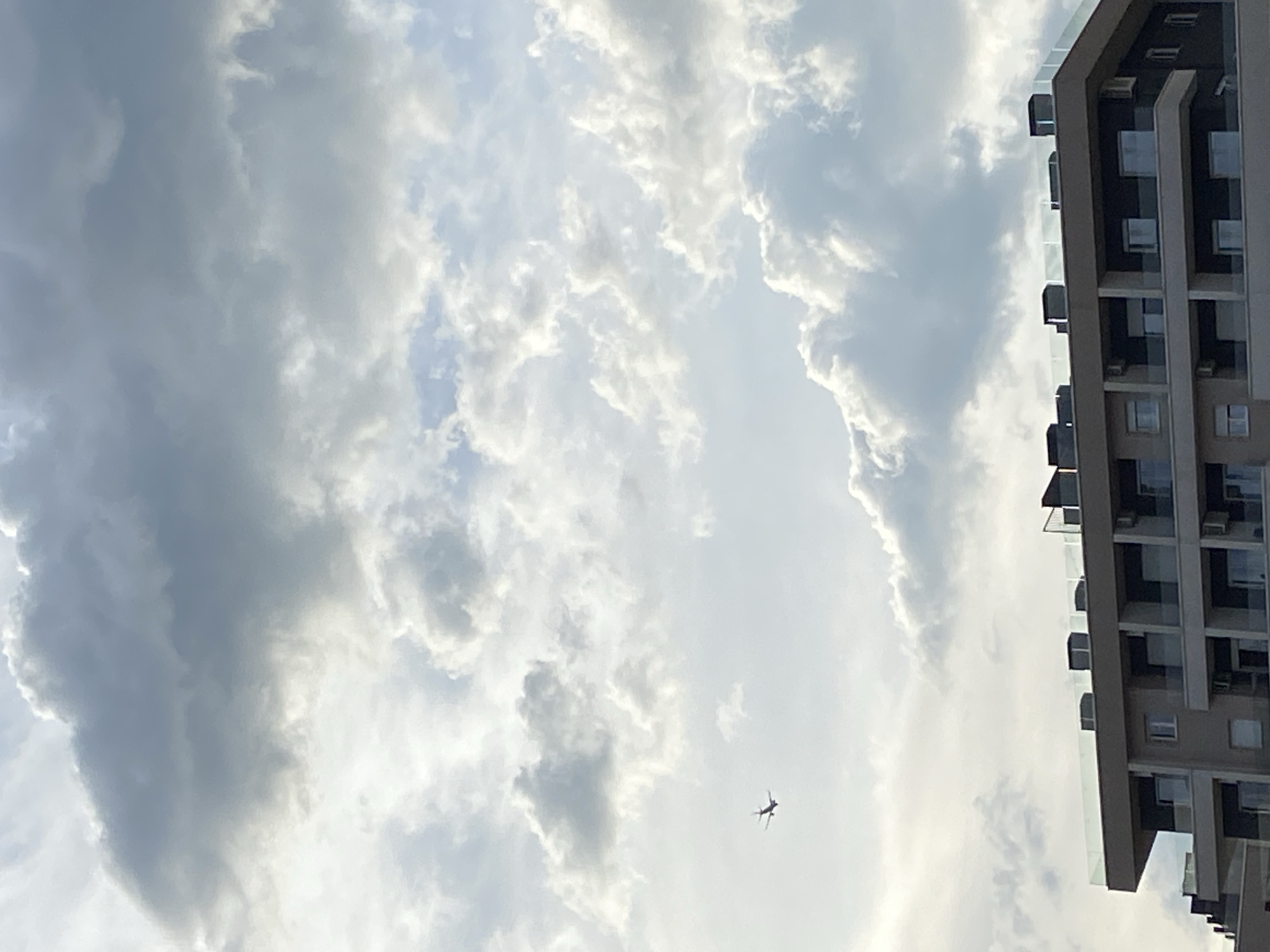 The image depicts a view of the sky with scattered clouds and a tall building on the right side. A small airplane is visible in the lower center of the frame. The building appears to have multiple floors with windows and balconies. Given that I have no specific landmarks to pinpoint, I am unable to confirm the precise location in Budva, Montenegro with only this image.