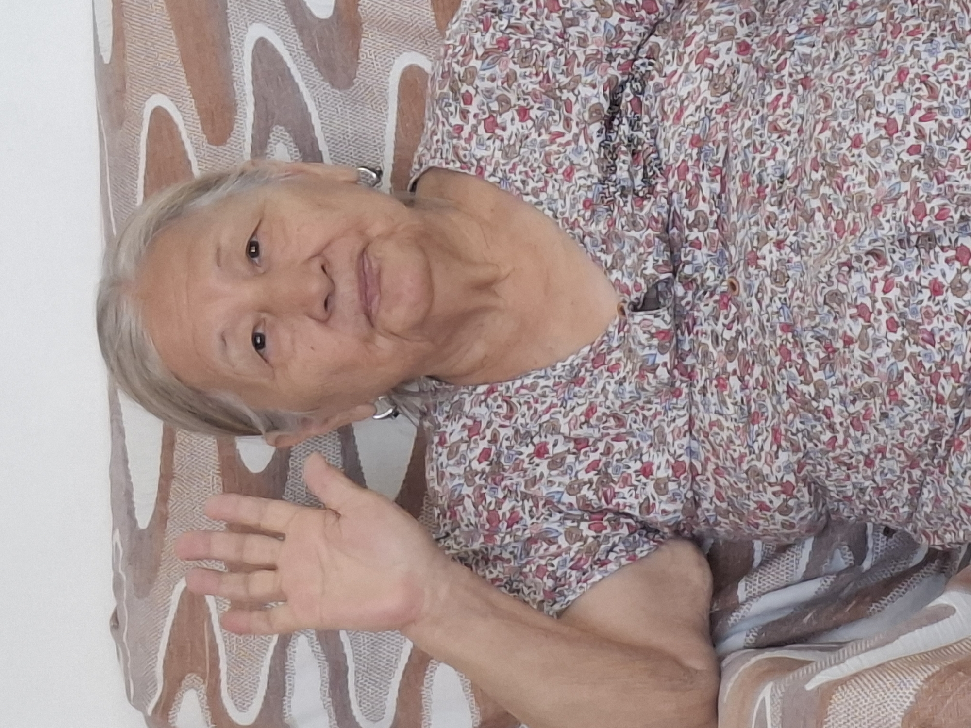 A close-up, medium shot captures an elderly woman with gray hair, her face positioned slightly off-center. She is seated and facing forward, with her right arm raised and her hand open, fingers splayed. She wears a floral print shirt with a V-neck and small buttons. The background features a patterned sofa and a plain wall. The image appears to have been taken indoors. There is no indication of the specific location in the city of Almaty, Kazakhstan.