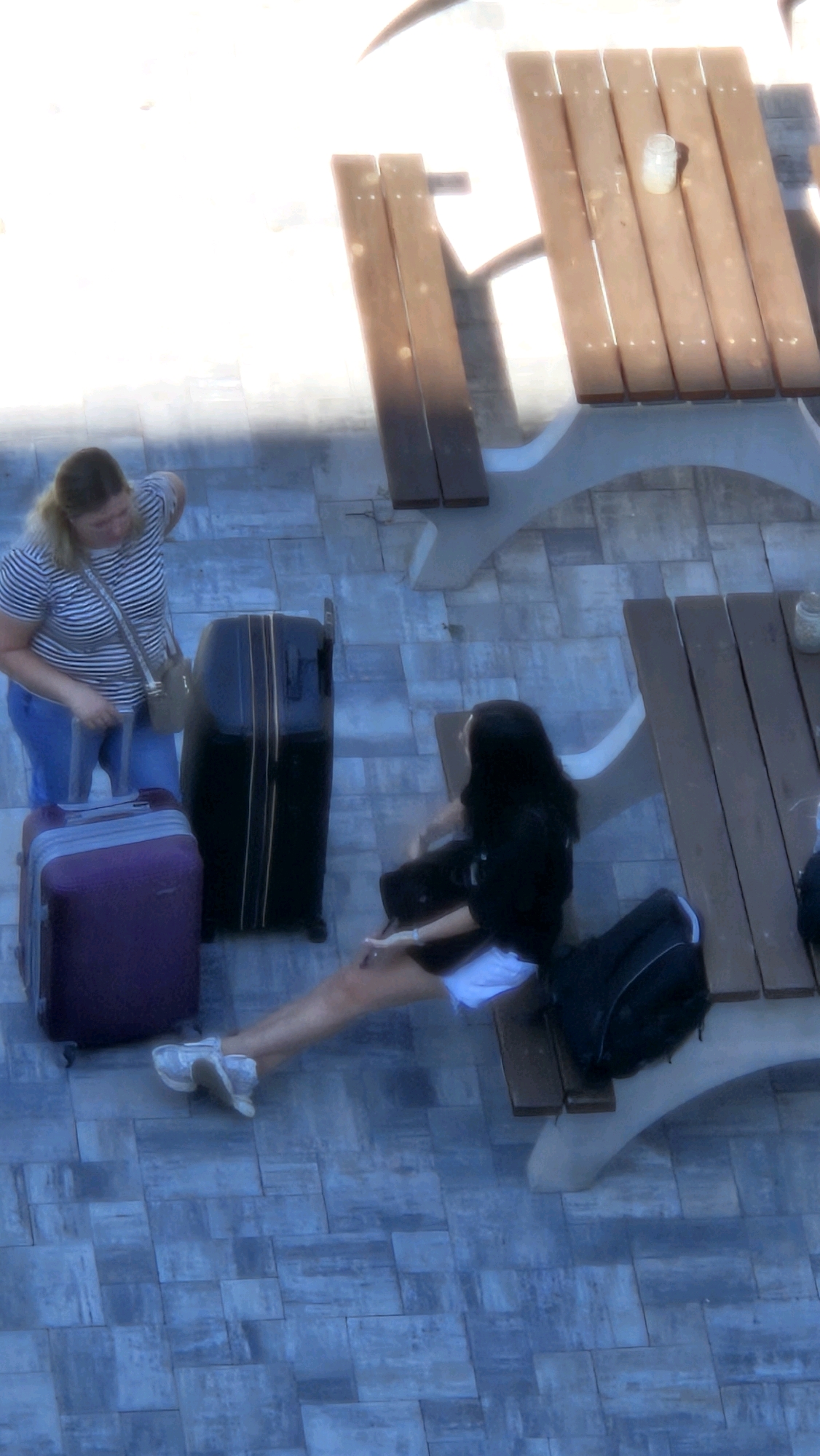 A woman with blonde hair, wearing a striped t-shirt and blue pants, stands next to a purple suitcase and a large black suitcase. She is holding the handle of the purple suitcase. To her right, another person with long dark hair, wearing a black hoodie and white shorts, is sitting on a bench next to a picnic table. This picnic table is made of wood and has a clear glass on it. The scene appears to be an outdoor patio area paved with grey rectangular tiles. The image does not provide any specific visual cues to identify the city as Prešov, Slovakia.
