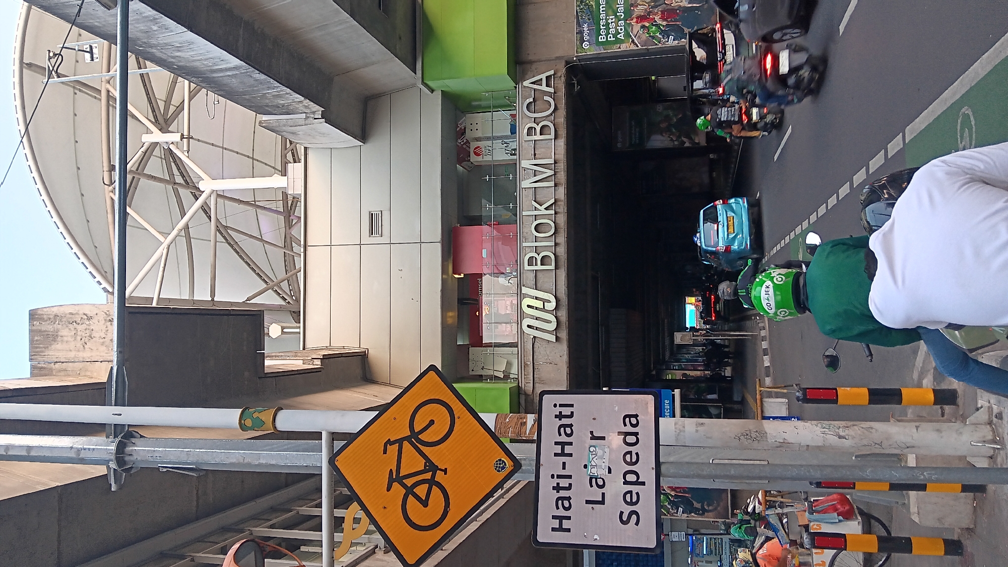 An eye-level, full shot captures a street scene under an elevated structure in South Jakarta, Indonesia. Dominating the foreground is a street sign with a yellow diamond shape and a black bicycle symbol. Below it, another sign reads "Hati-Hati Lajur Sepeda," indicating caution for bicycle lanes. A traffic light pole stands to the left, adorned with a green, yellow, and black decorative band.
The elevated structure overhead appears to be part of a transportation hub, possibly a train or bus station, with a white, curved, translucent roof supported by a white framework. The buildings beneath the structure have large windows and storefronts, with a prominent sign reading "Blok M BCA."
The street is a multi-lane road with visible traffic. Several motorcycles are present, some with riders wearing green jackets and helmets, one of which clearly displays the "GOJEK" logo. A blue car is also visible on the road. Pedestrians are seen on sidewalks and within the covered areas of the infrastructure. The overall environment suggests an urban setting with active transportation and commercial activity. The lighting indicates it is daytime, possibly late afternoon, given the angle of the sun.