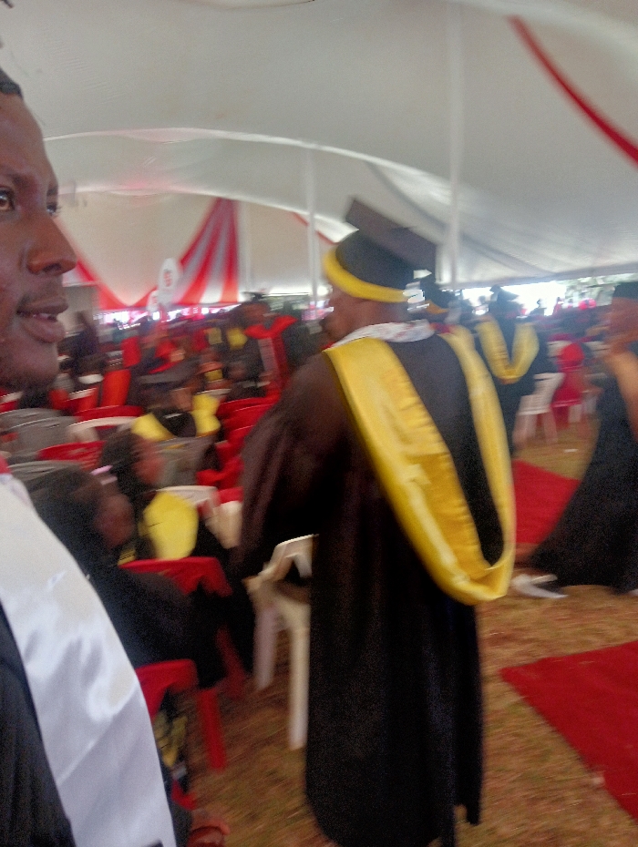 The scene depicts an outdoor event taking place under a large white tent with red decorative fabric accents, located in Shamenei, Kenya. The ground surface consists of dry, light-brown earth, partially covered by red carpets. In the immediate left foreground, the partial profile of a dark-skinned male is visible, facing right, wearing a white academic stole over a dark garment. Centered in the midground, an individual is observed from behind, attired in a black academic gown, a prominent yellow academic hood, and a black mortarboard cap with a yellow band. Numerous other dark-skinned individuals are present in the background, many wearing similar academic gowns and hoods (some yellow, some red), seated on red and white plastic chairs, or standing. The overall environment indicates a formal gathering or ceremony.